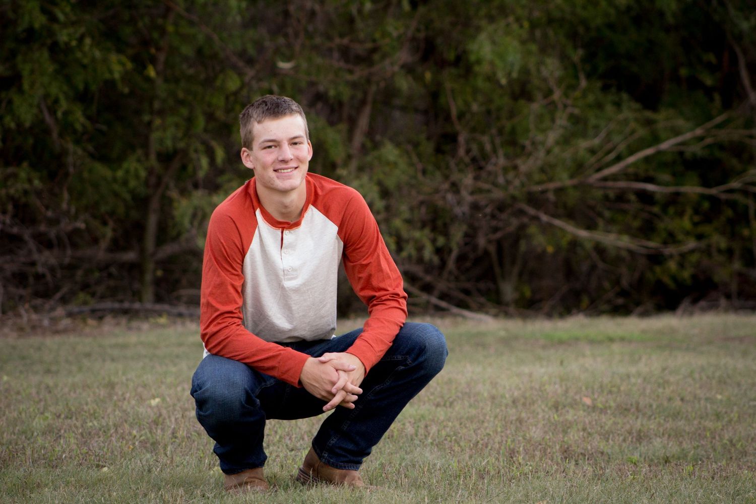 Teenage boy in red and white shirt crouching and smiling outdoors on green grass.