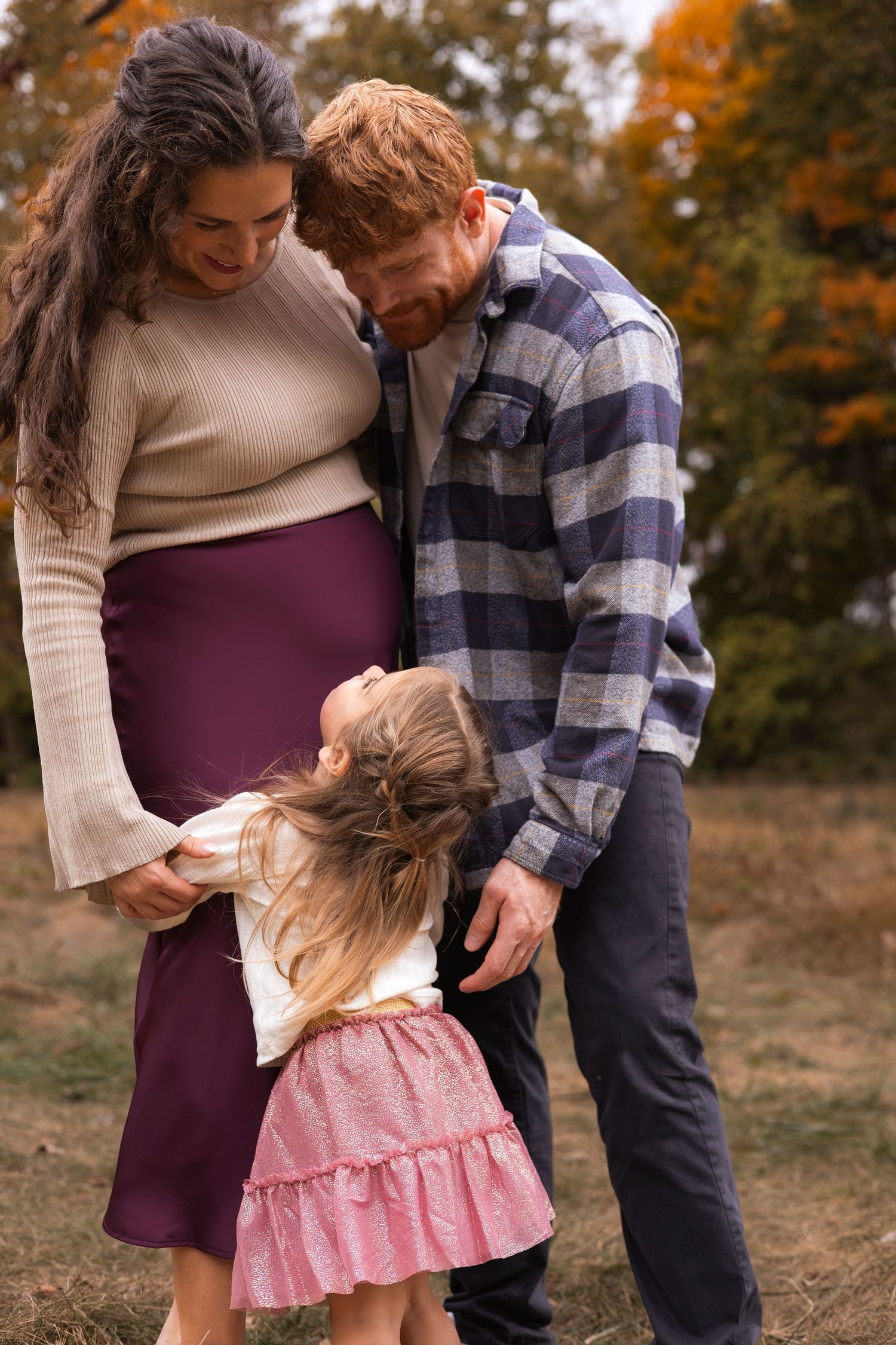 Red-haired father and pregnant mother smile as their young daughter kisses moms baby bump in autumn park.