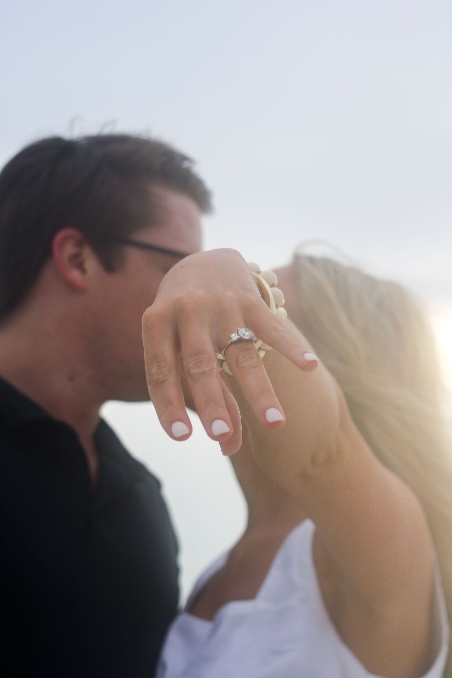 An engaged couple kissing while the woman displays her diamond engagement ring in bright sunlight.