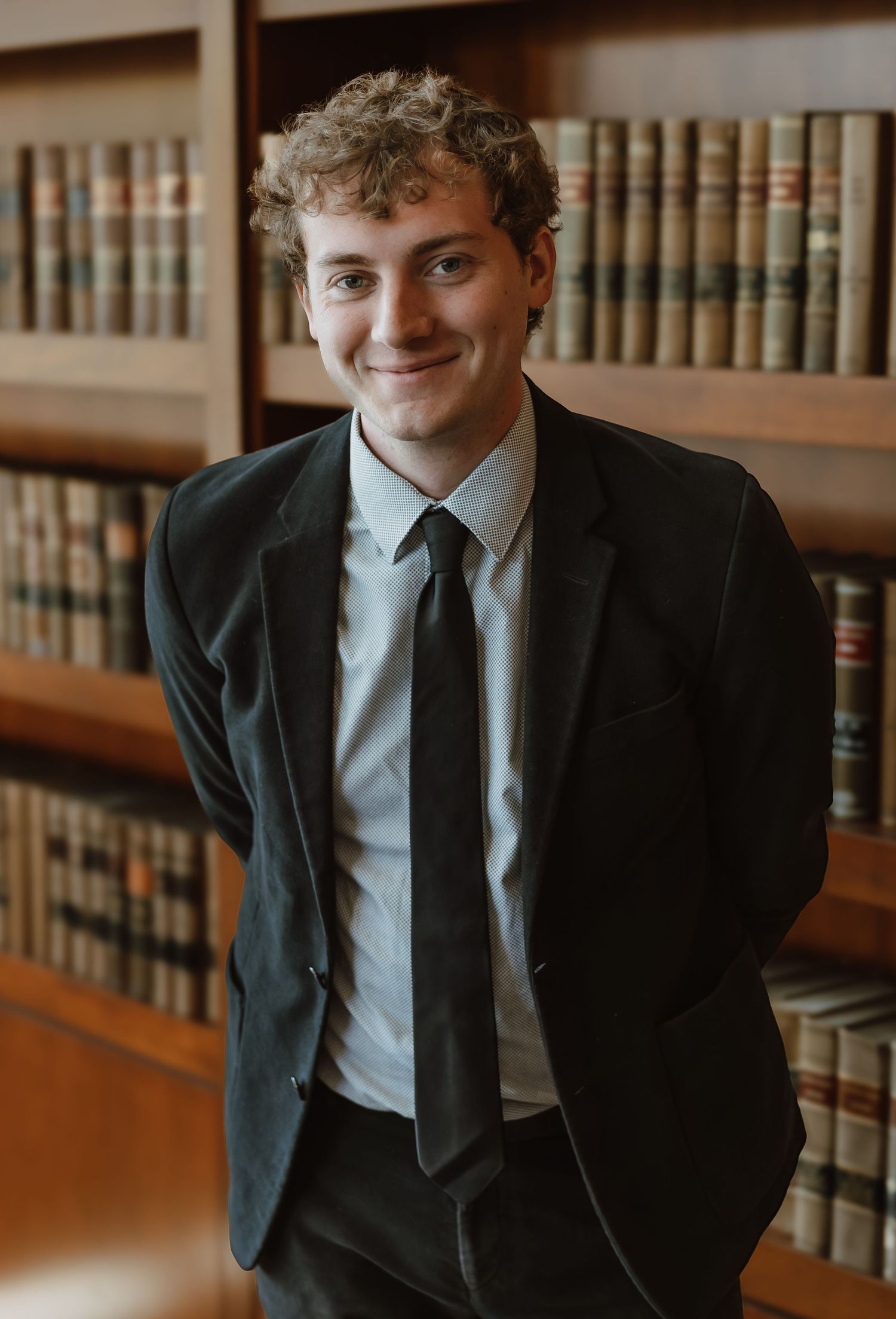 Young man in black suit and tie smiling in front of law library bookshelves.