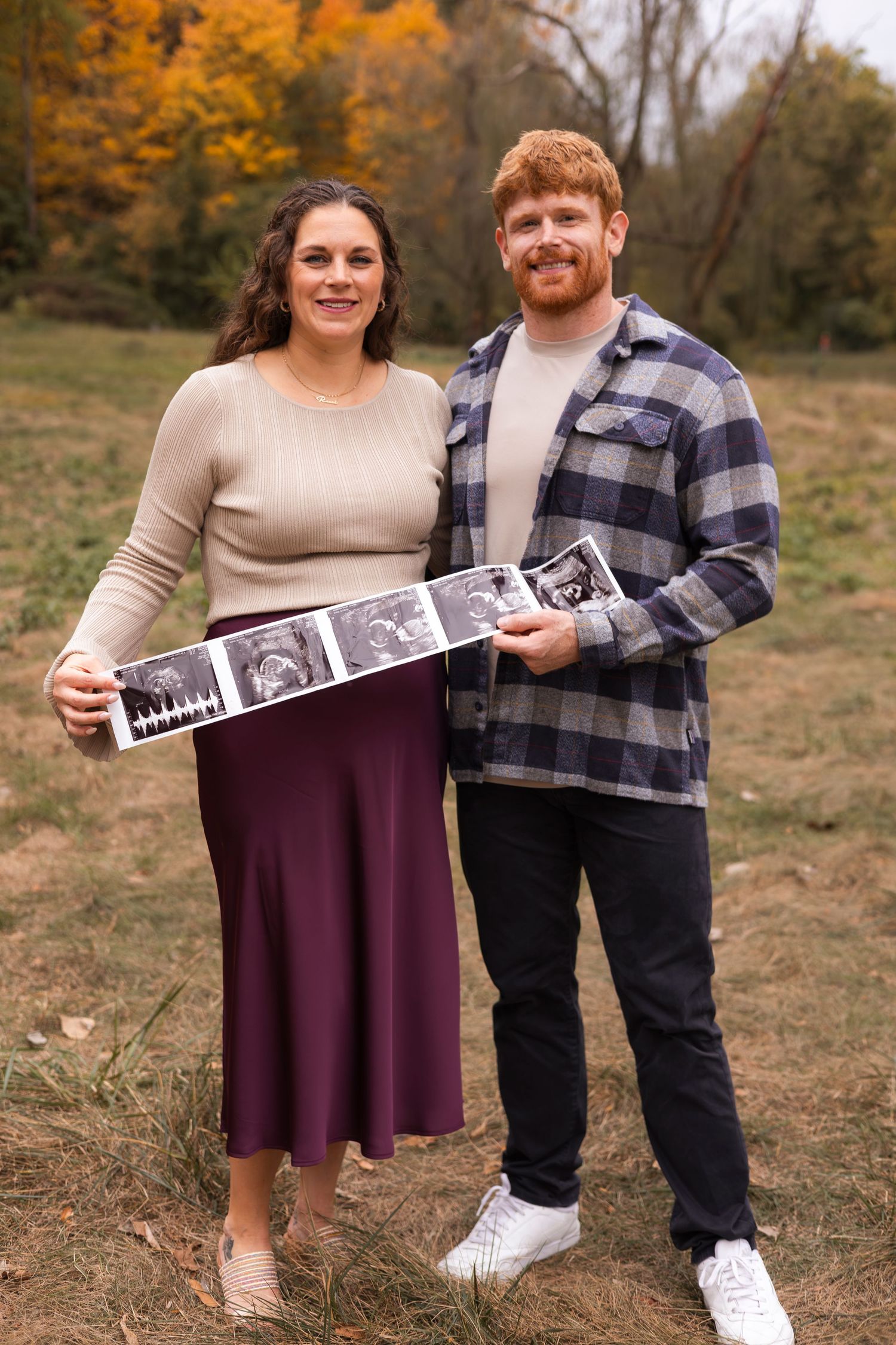 Expecting couple holding ultrasound photos in an autumn field, woman in burgundy skirt and man in plaid shirt smiling.