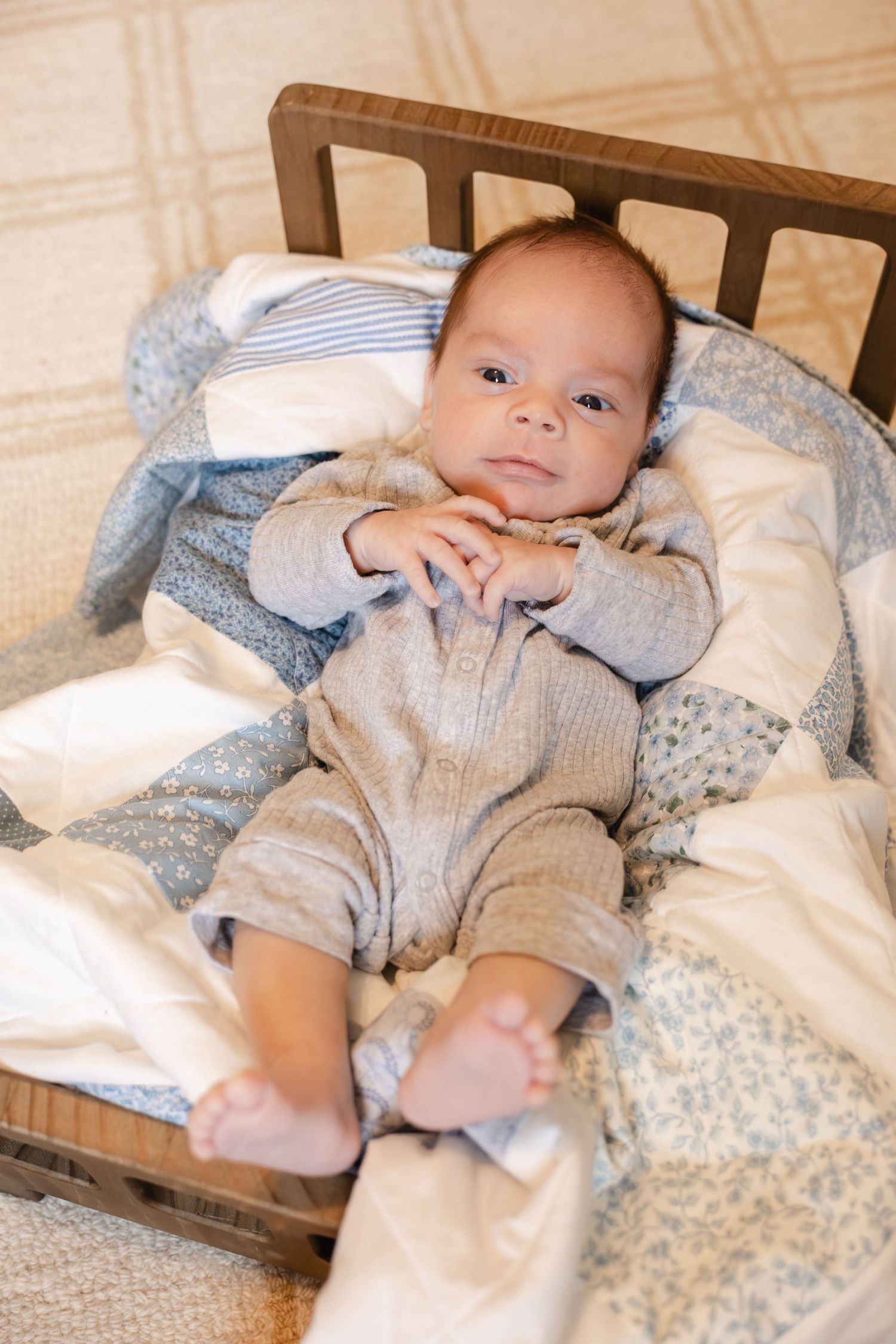 Sweet newborn baby in gray footie pajamas lying in small wooden bed with blue blanket.