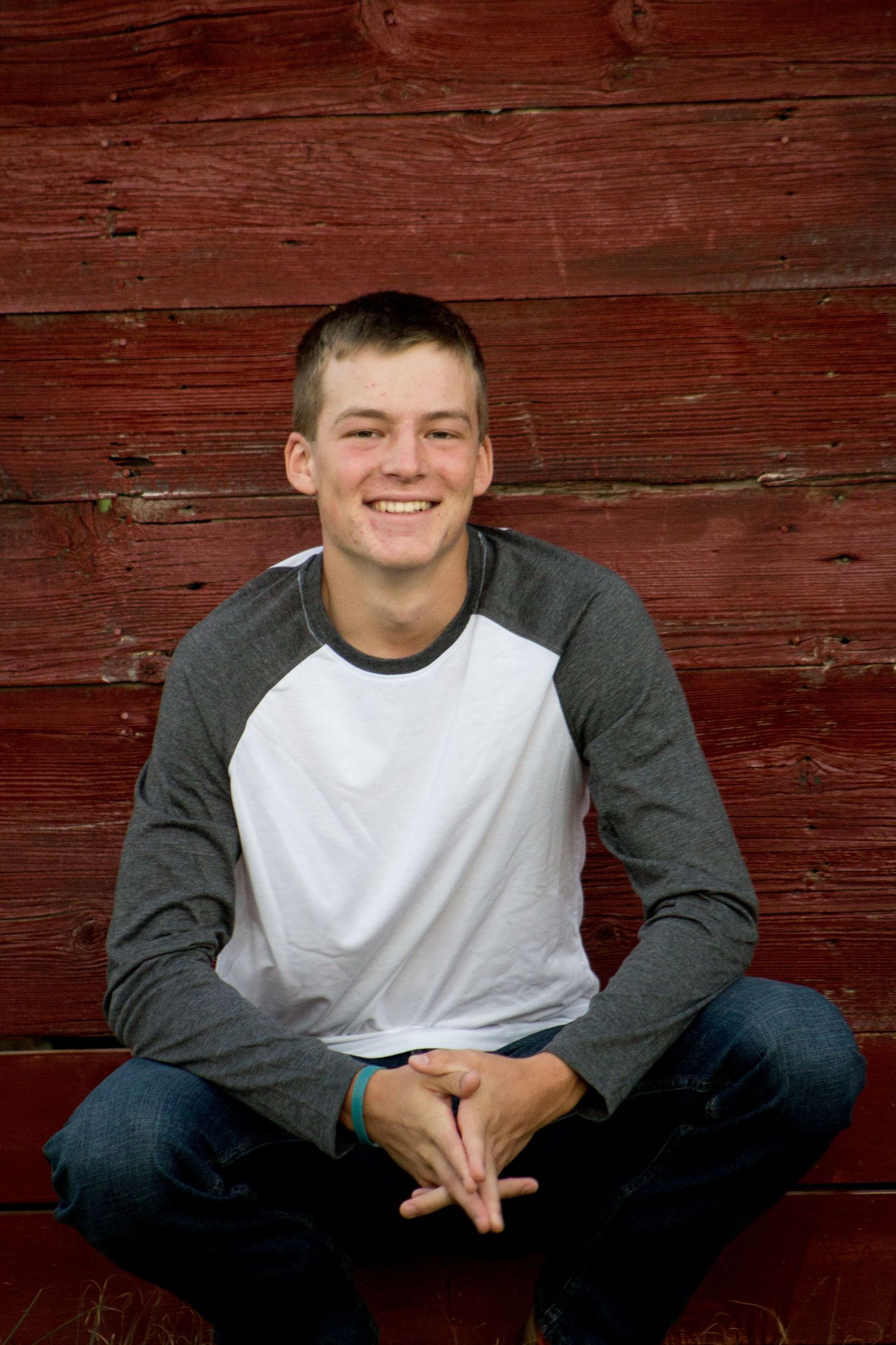 Smiling teenage boy sitting against rustic red barn wall for senior portrait.
