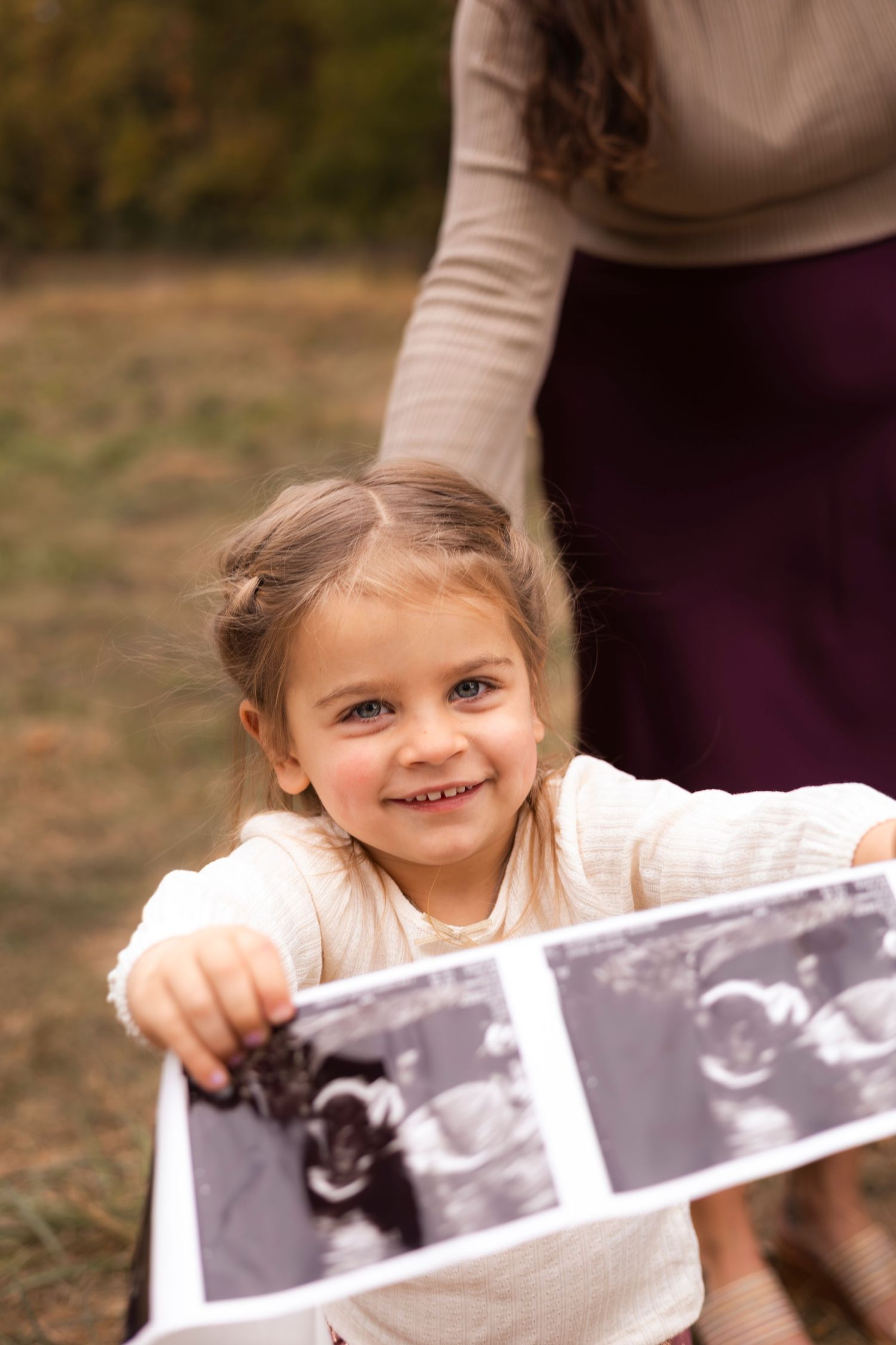 Happy young girl proudly displays ultrasound photos toward camera during fall maternity photo session outdoors.