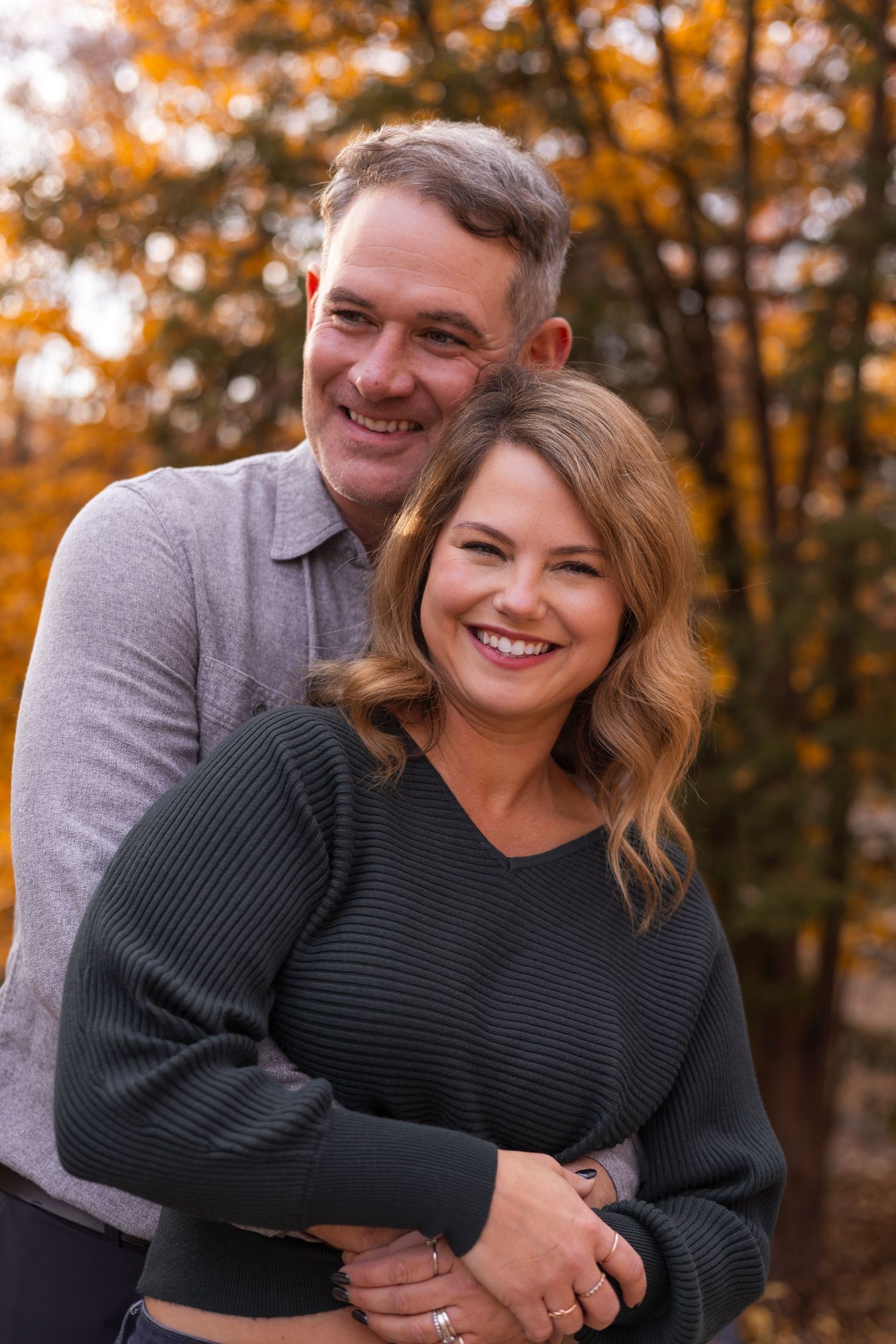 Smiling couple posing together outdoors with vibrant autumn trees behind them.