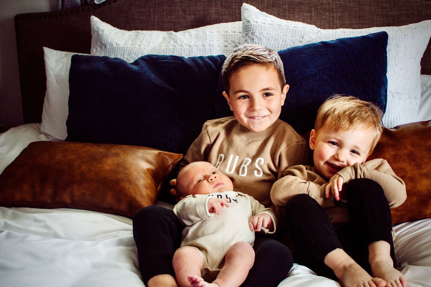 Three children sit together on a white bed, the eldest holding a newborn while a toddler smiles warmly.