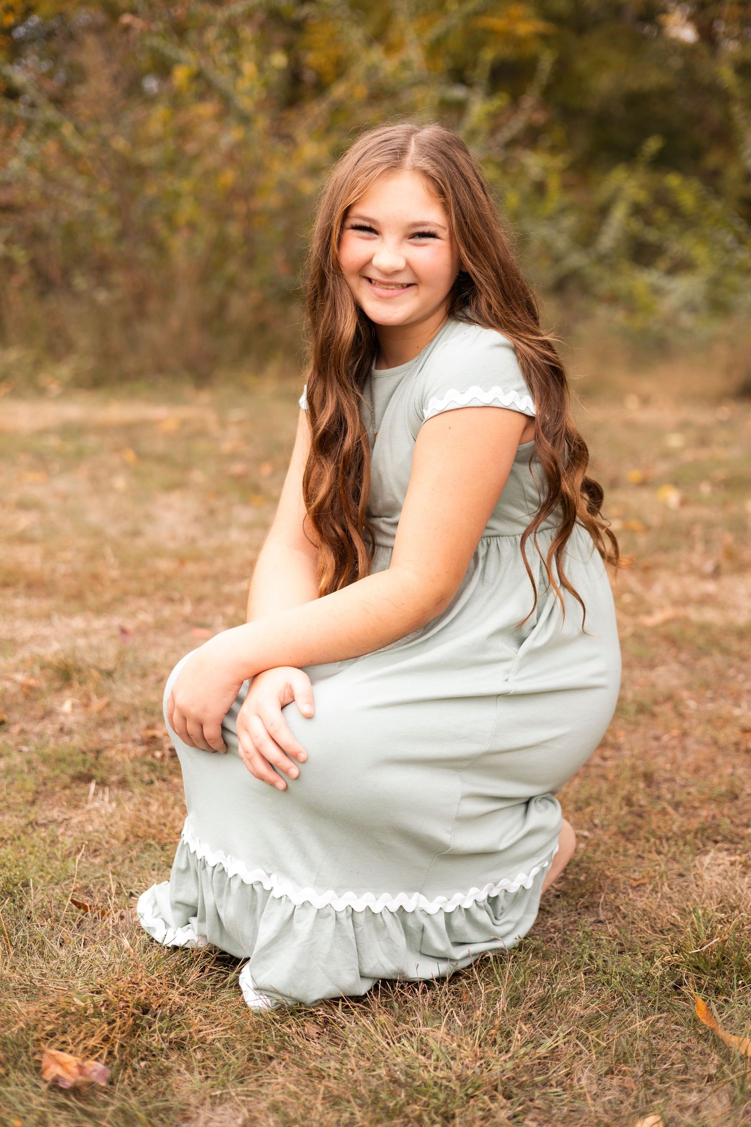 Young girl in sage green dress crouching outdoors for fall portrait session.