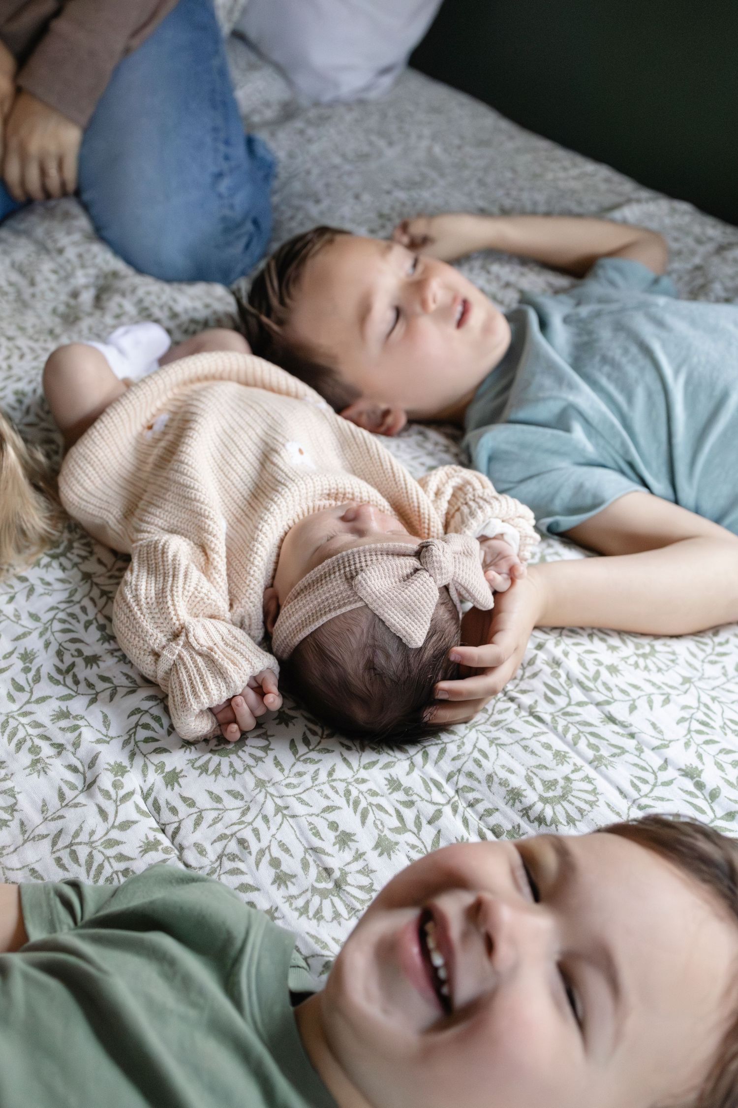 Two children lay on a floral bed with a sleeping newborn baby girl in a pink bow headband.
