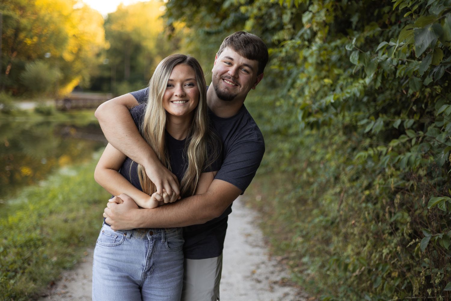 A happy young couple hugging closely on a scenic outdoor path lined with green foliage.
