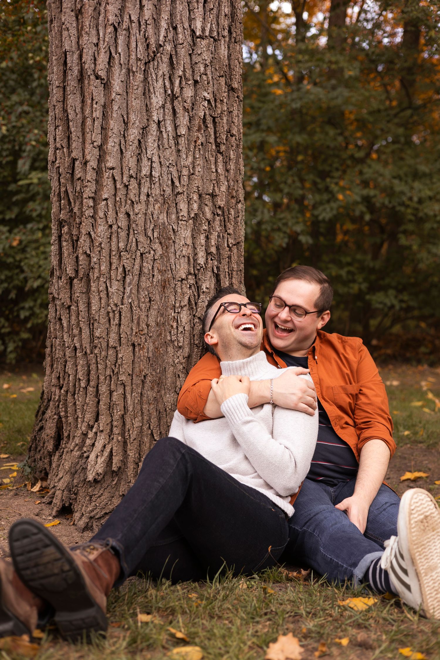 Two men sitting against a tree trunk laughing in an autumn park.