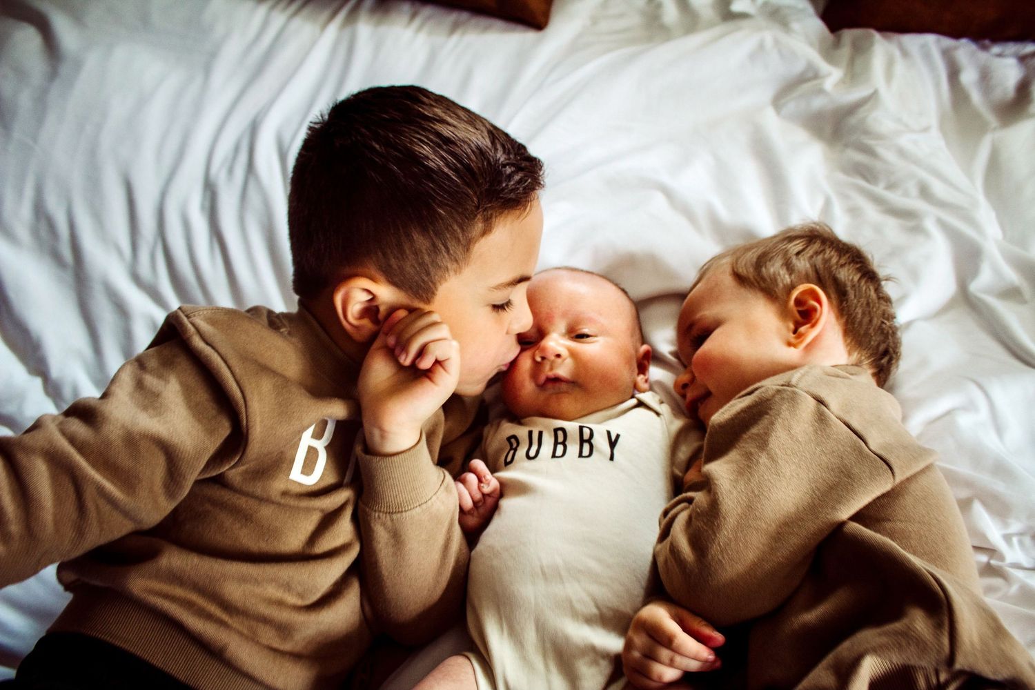 Two older brothers lean in to kiss their newborn sibling wearing a 'Bubby' onesie on a white bed.