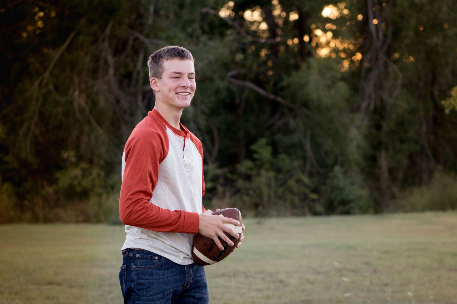 Smiling teenage boy in red and white jersey holding a football in an outdoor field.