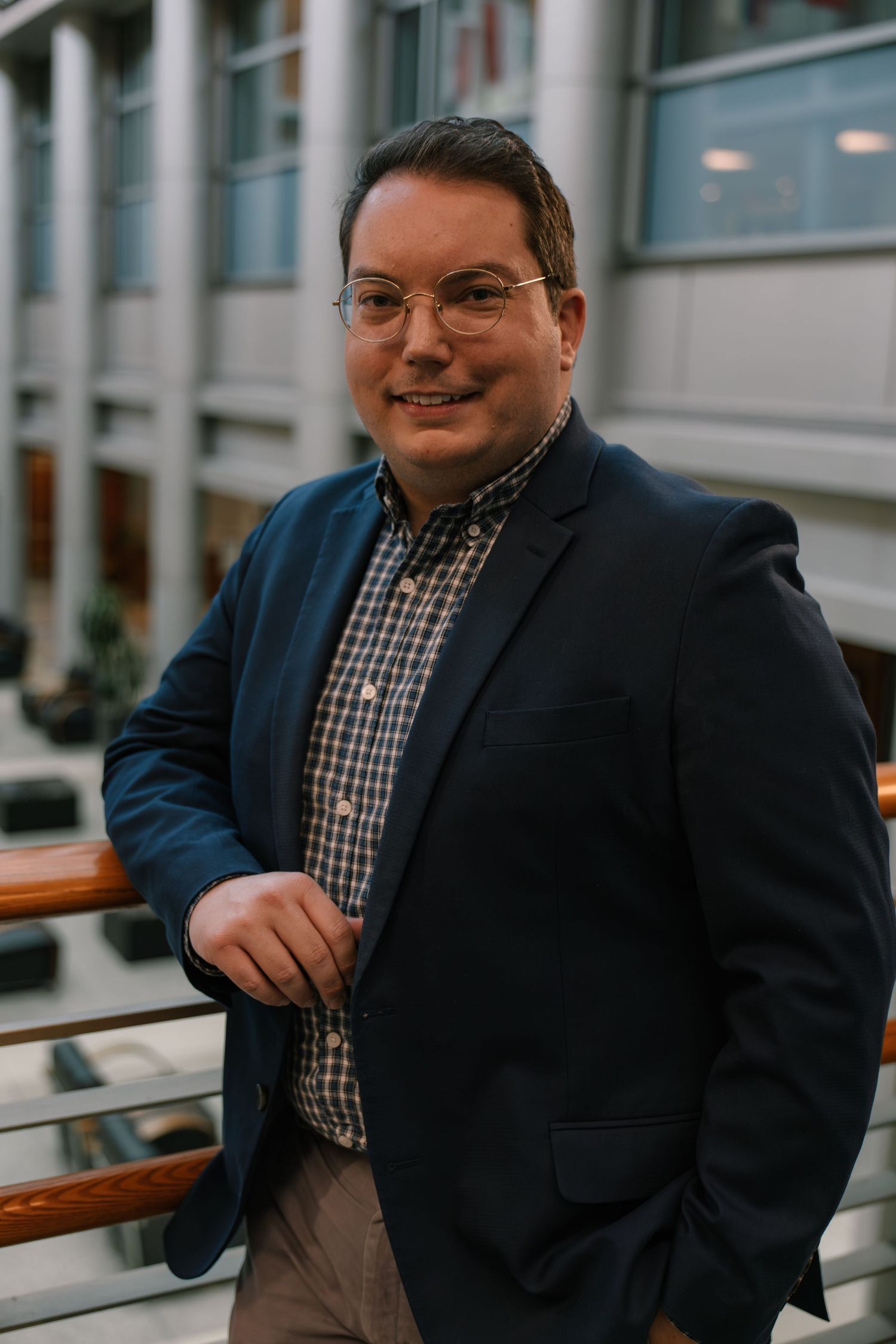 Man in blue blazer and glasses smiling while leaning on railing in modern building atrium.