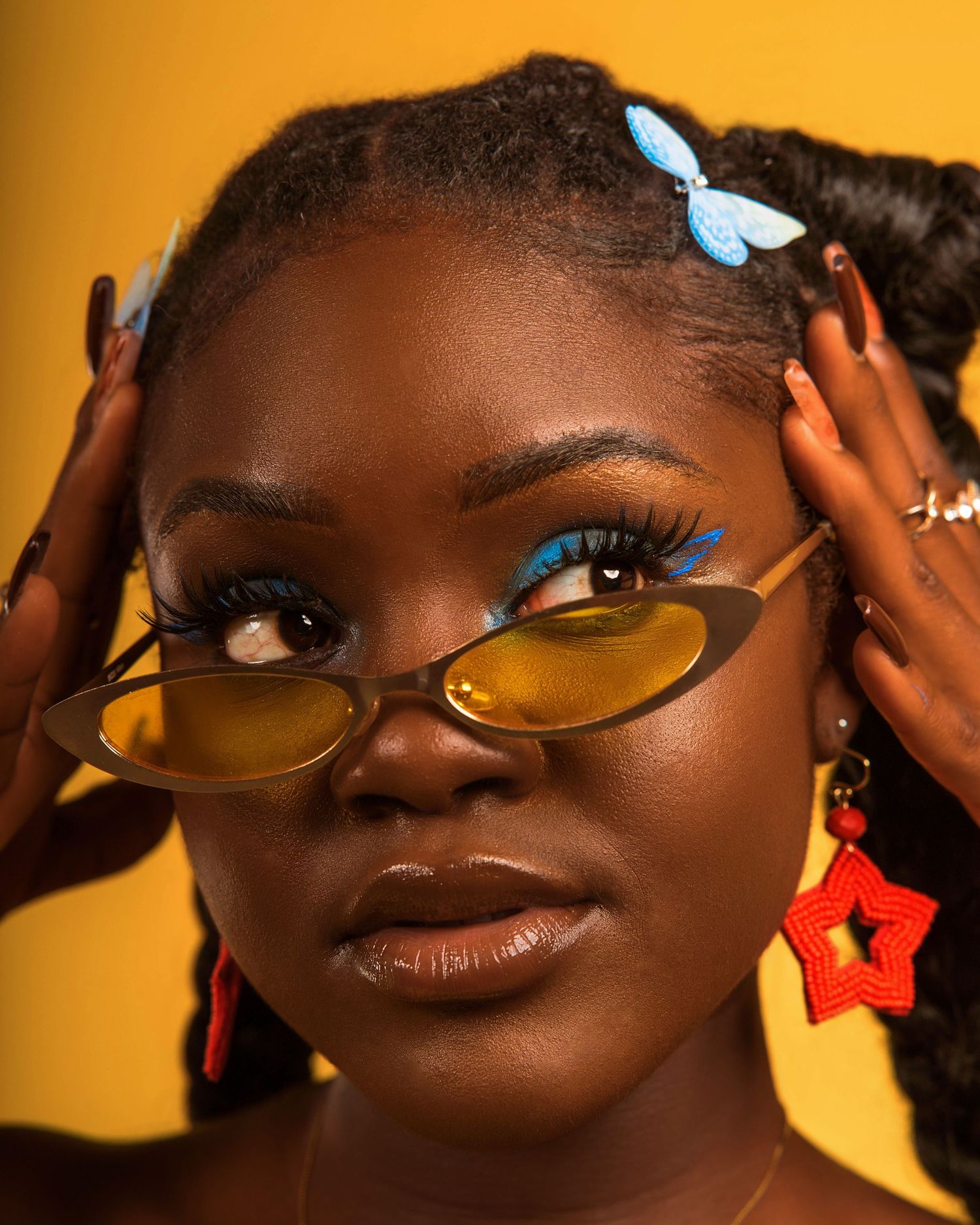 Artistic portrait with tinted glasses and butterfly hair clip against orange background.