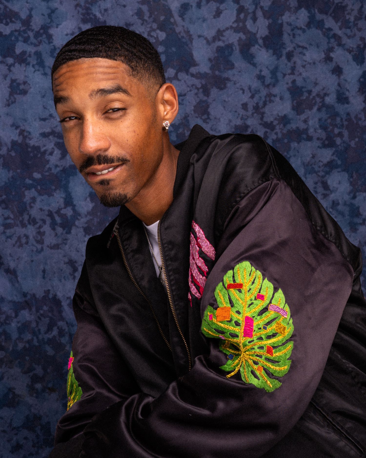 Young man in black satin bomber jacket with tropical leaf embroidery poses against blue textured background.