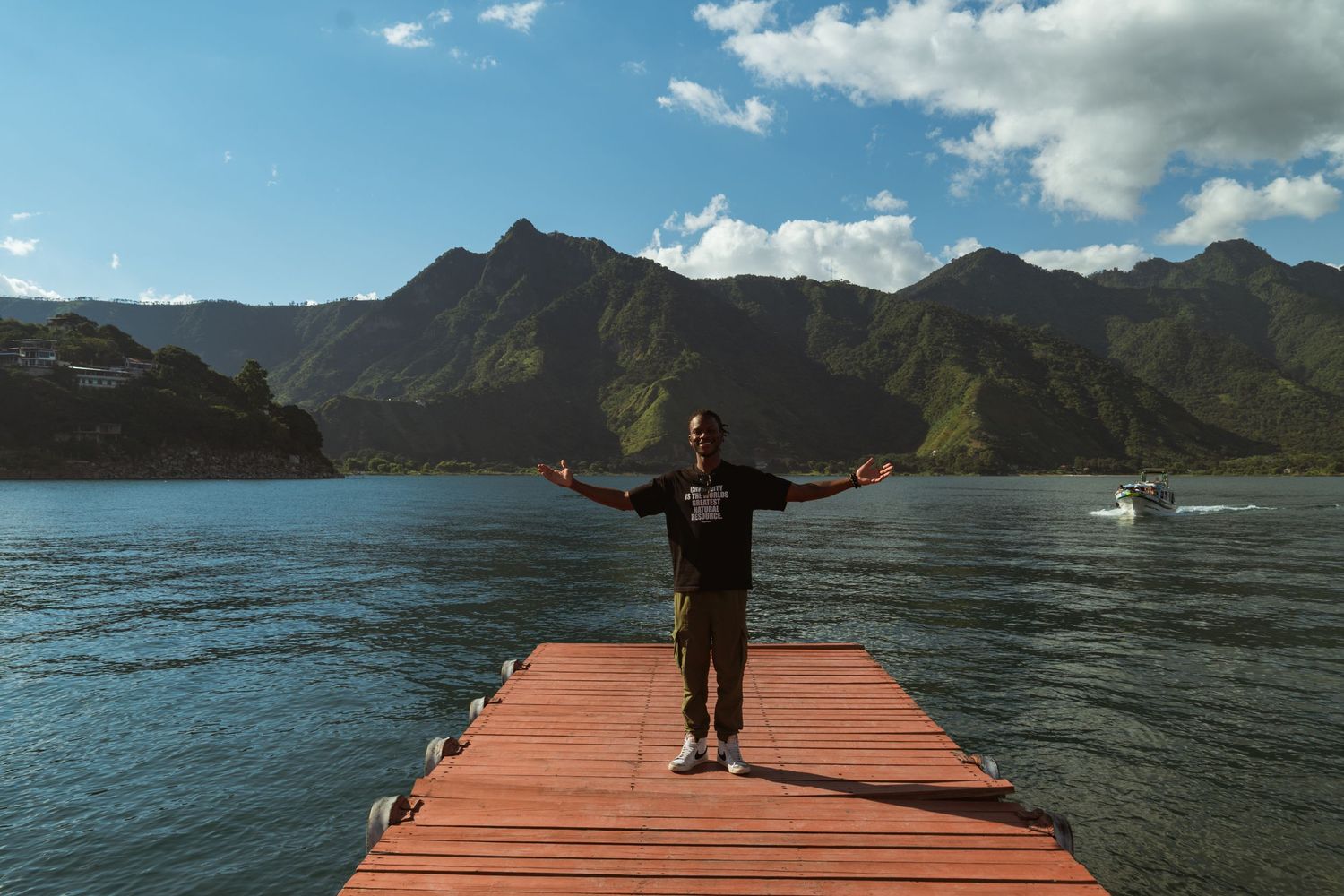 A figure stands with outstretched arms at the end of a wooden pier overlooking a scenic mountain lake on a cloudy day in Lake Atitlan.