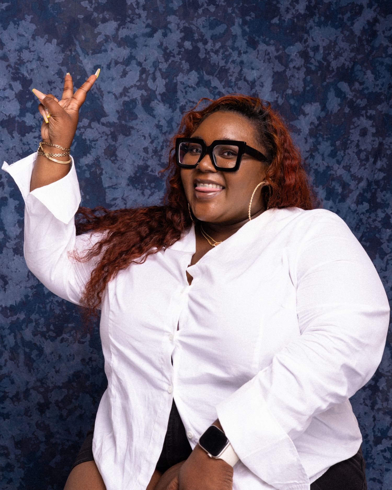 Woman in white shirt and black glasses smiling and posing with hand gesture against blue backdrop.