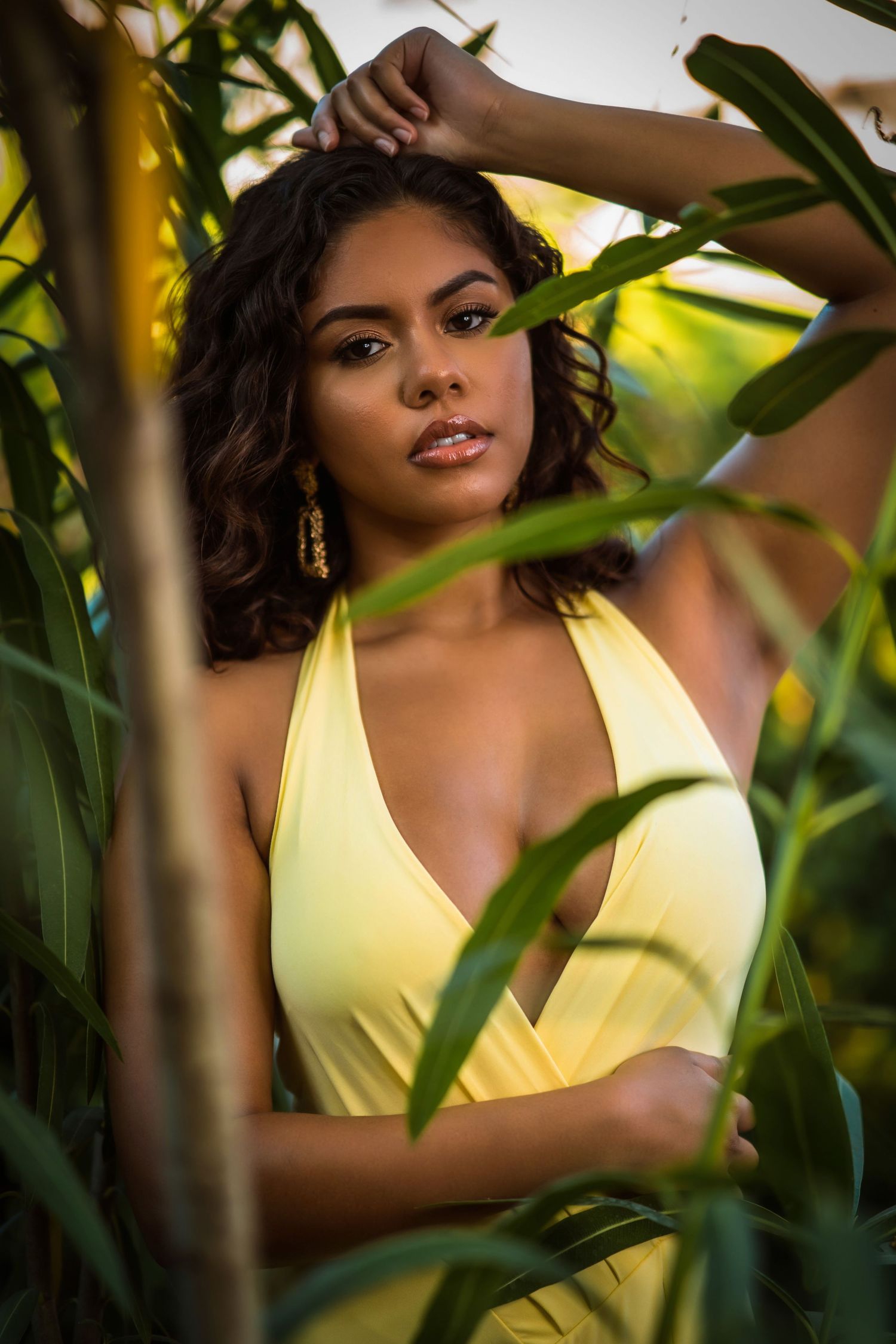 Young woman in yellow halter dress posing among tropical green plants, wearing gold earrings and nose ring.
