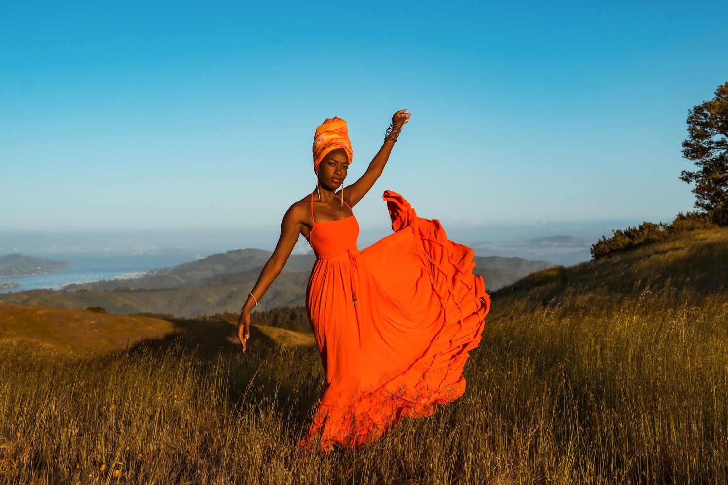 Silhouette in flowing orange dress poses dramatically against mountain sunset landscape.