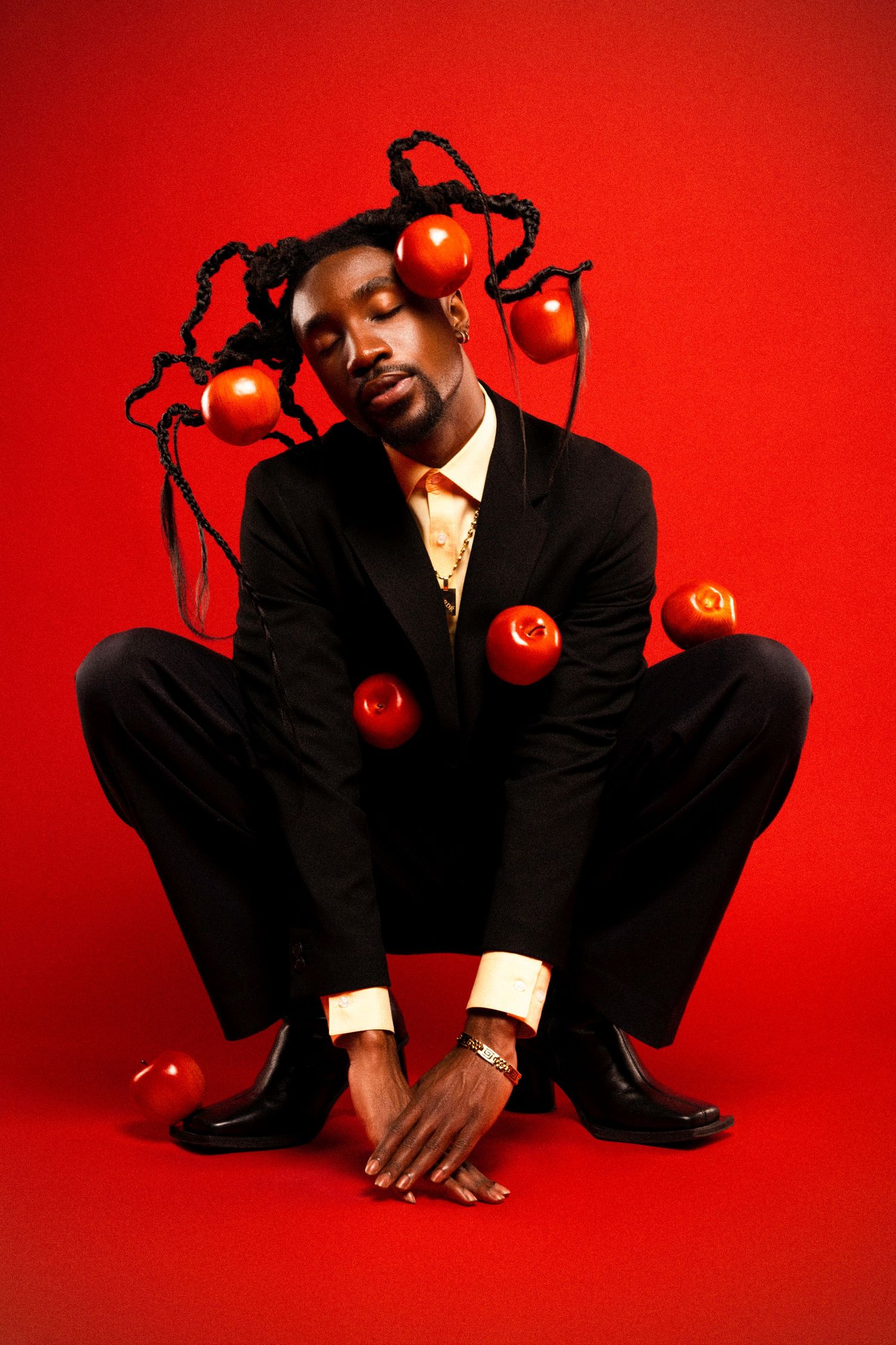 Fashion portrait with red apples suspended in braided hair while crouching in formal attire against red background.