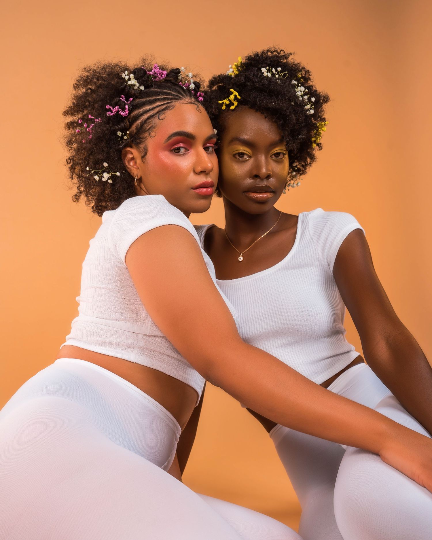 Two models in white outfits pose together for artistic portrait series against peach background.