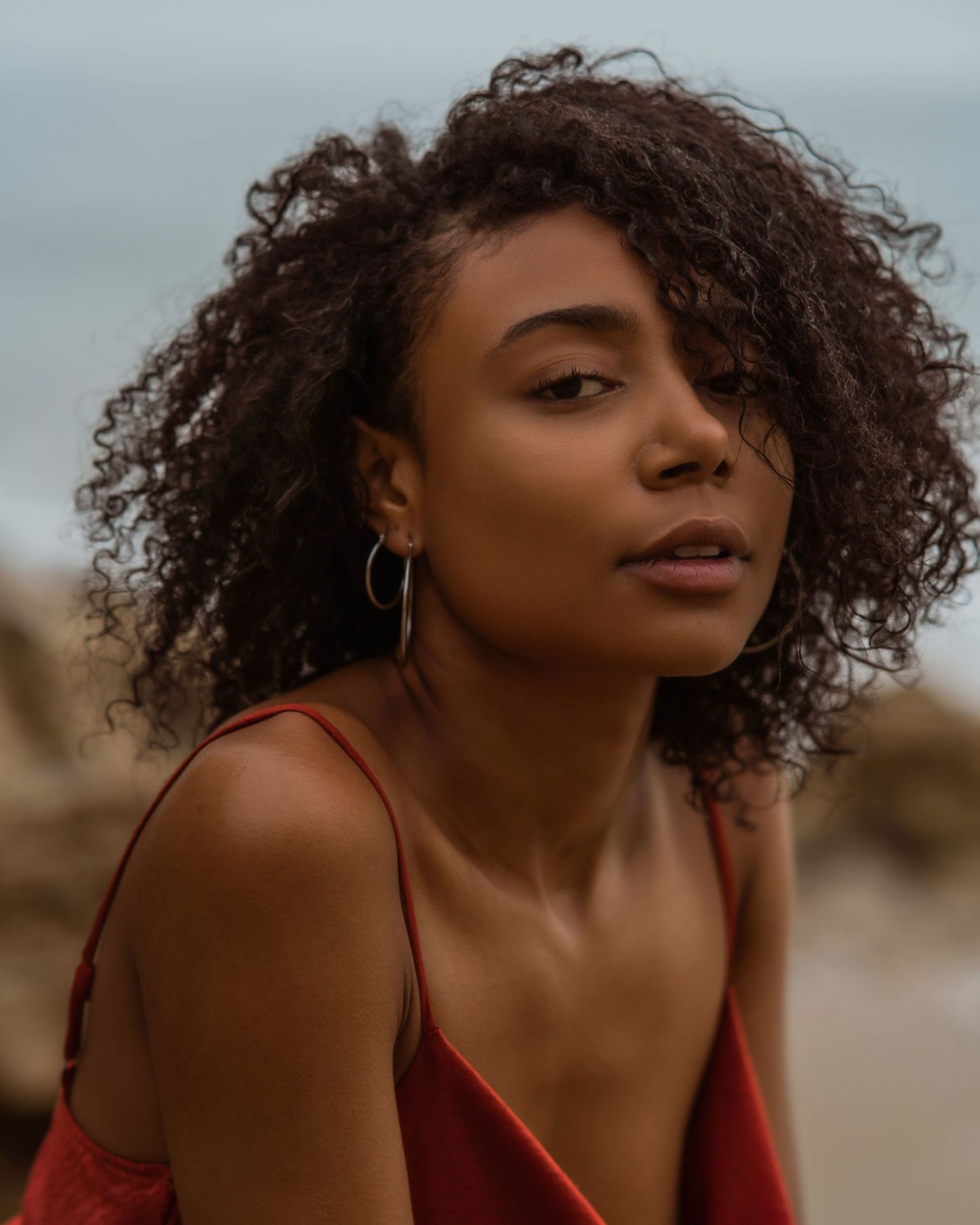 Young Black woman with curly hair wearing red top and hoop earrings poses at a rocky beach.