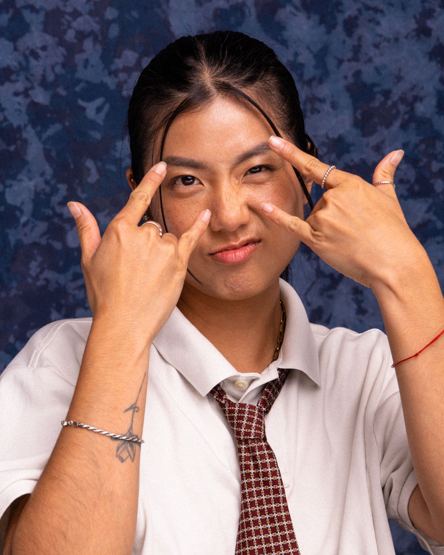 Young person in white shirt and tie making peace signs near eyes, posing against blue background.