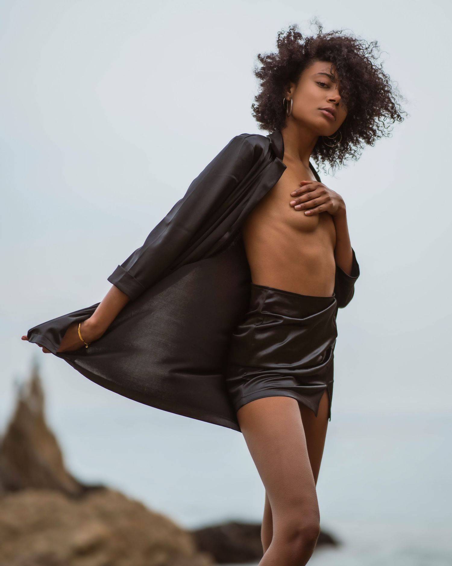 Woman in black satin skirt and open jacket poses on rocky shore with curly hair against overcast sky.
