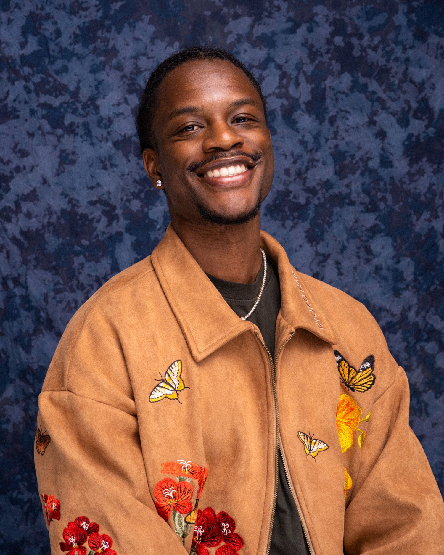 Smiling young man wearing a tan suede jacket with colorful butterfly and floral embroidery against a blue backdrop.