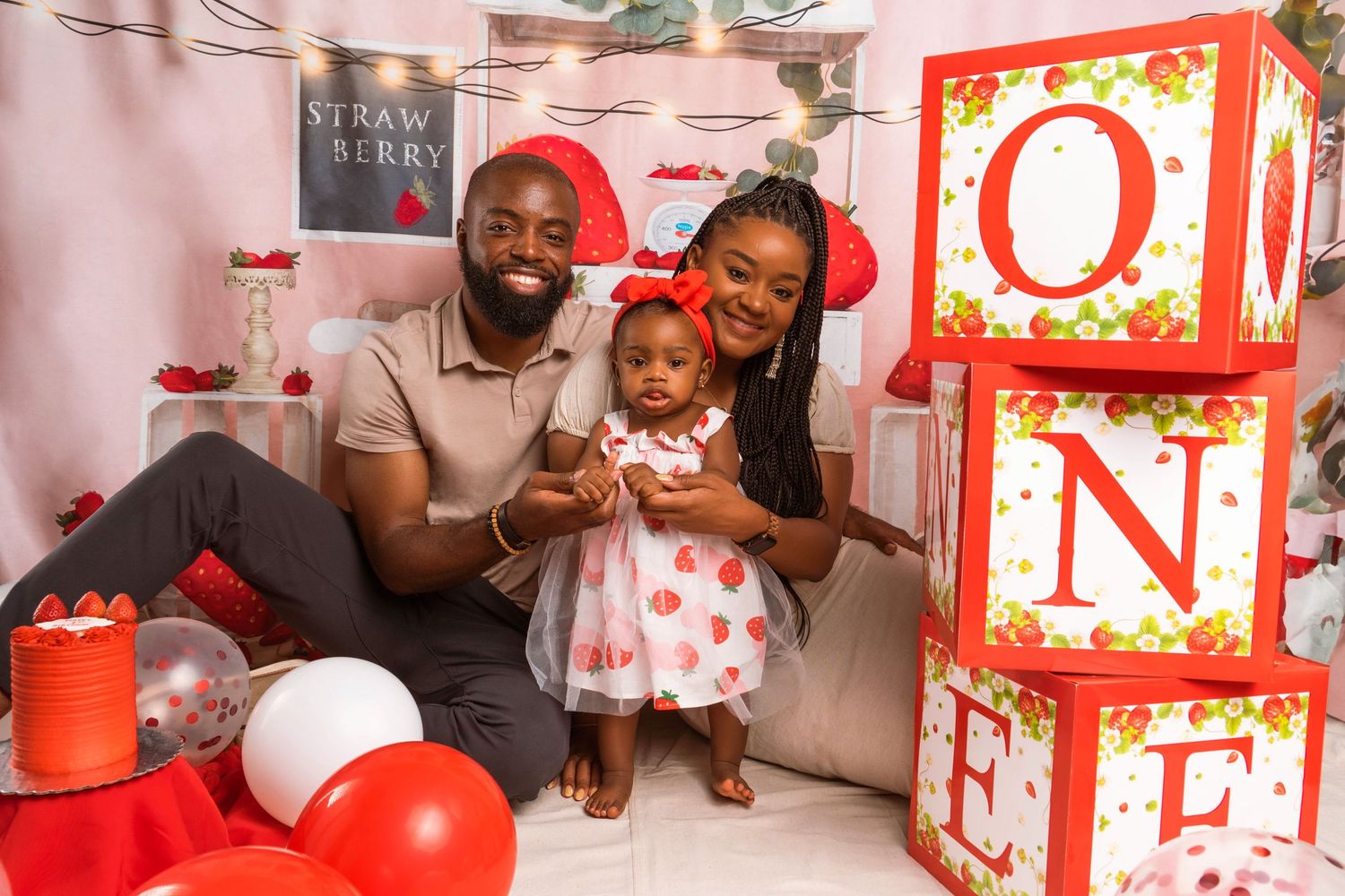 Family celebrating baby's first birthday with ONE blocks, red balloons, and strawberry theme decorations.