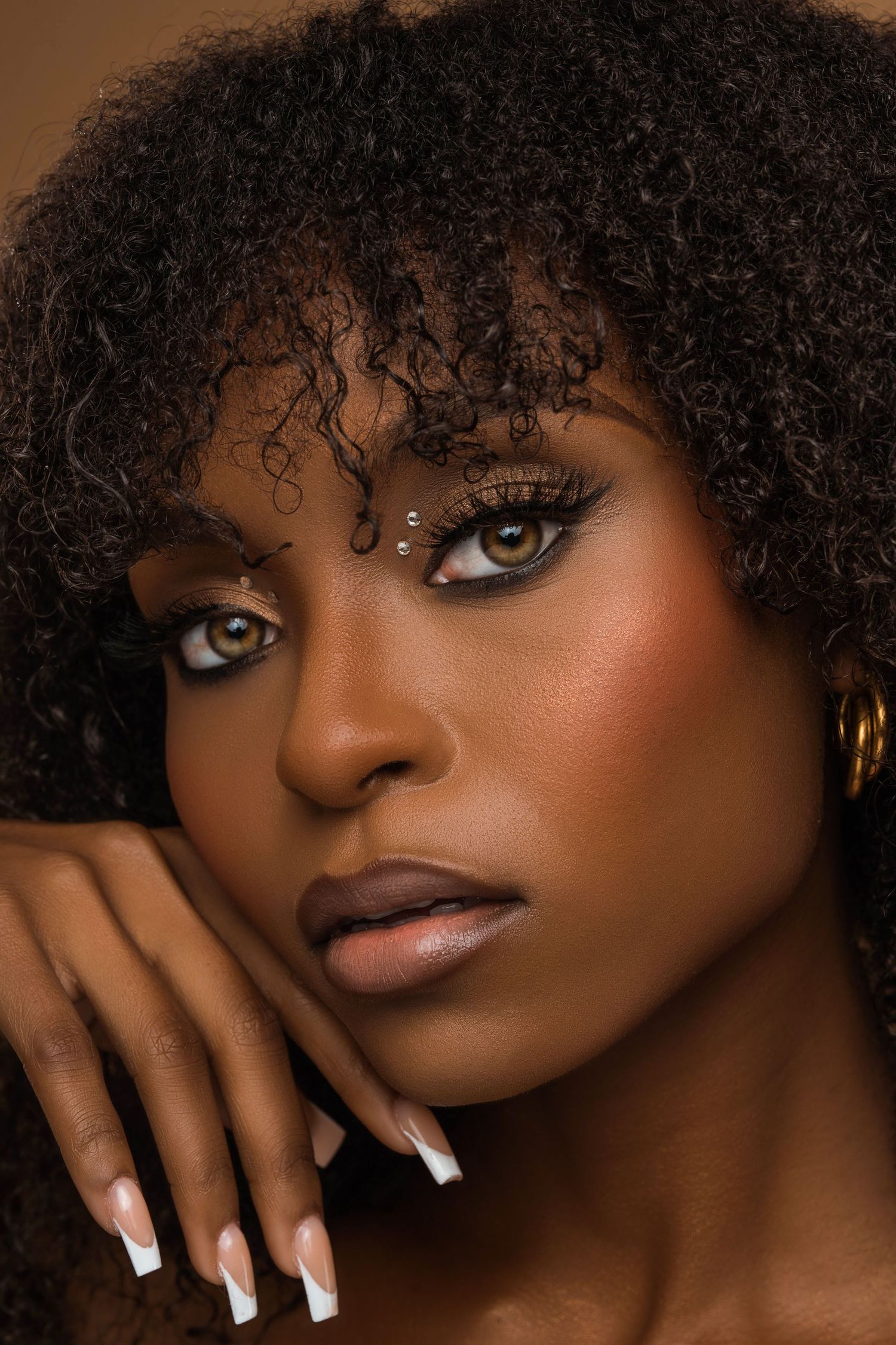 Beauty portrait with curly hair, dramatic makeup, and long white nails against a warm background.