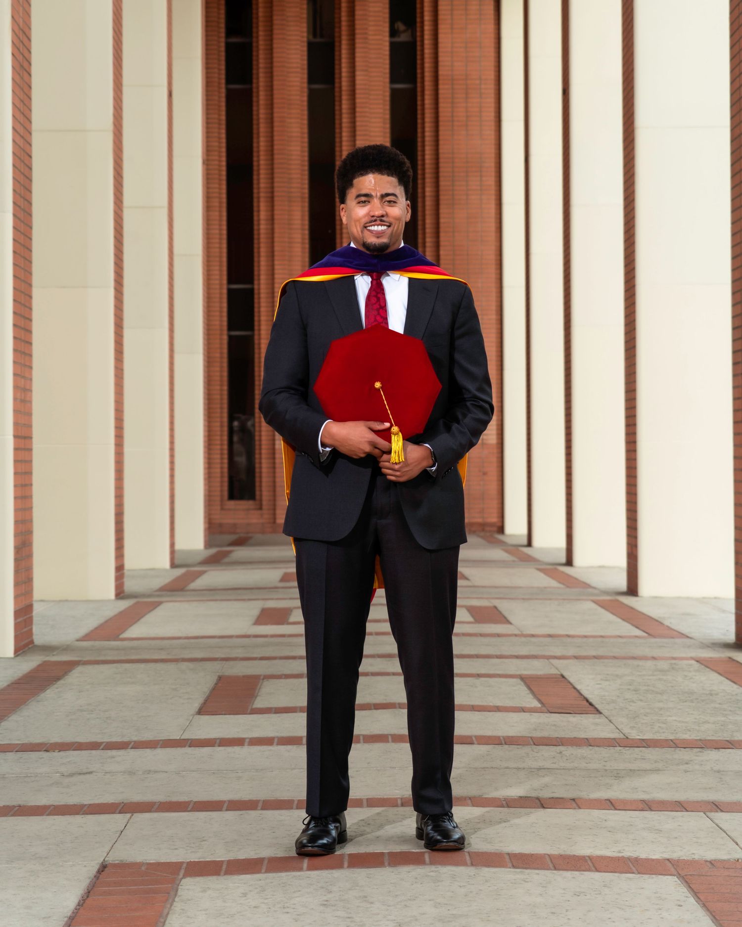 Graduate in formal attire standing in an architectural colonnade with striped pillars.