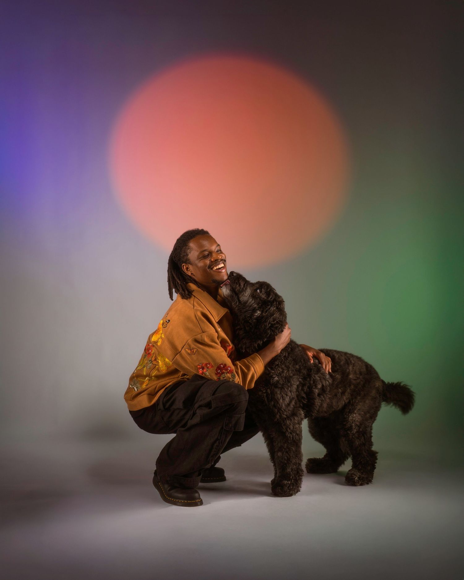 A playful interaction between a person in a brown jacket and a large black dog on a multi-color studio backdrop.