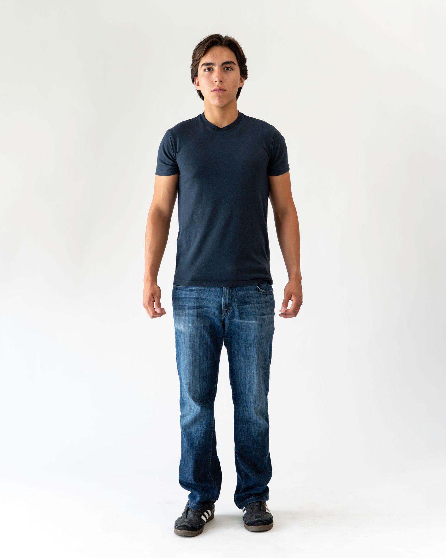 Man wearing a navy blue v-neck t-shirt and dark wash jeans, standing against a white background.