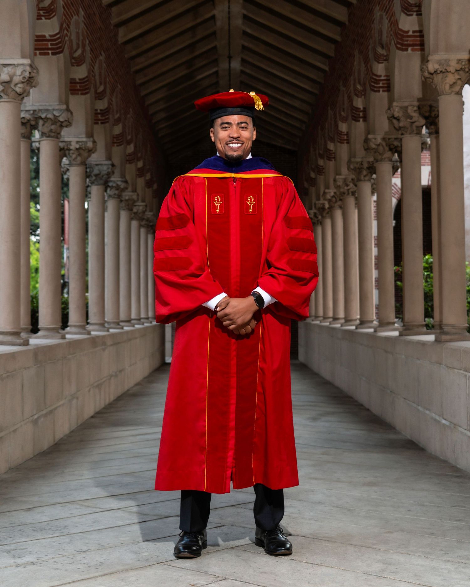 A graduate in a red cap and gown posing confidently in a historic architectural corridor during a milestone portrait photoshoot.
