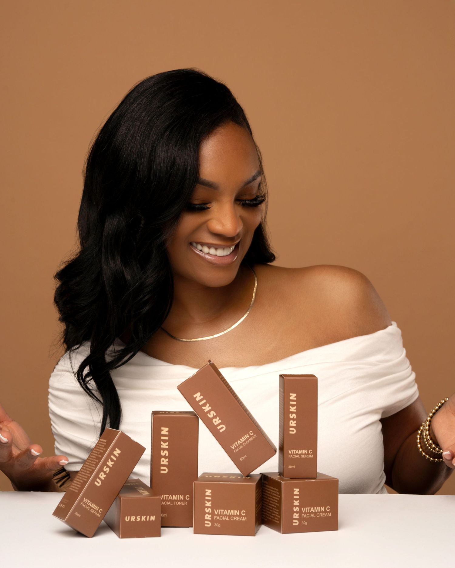 Smiling person in white off-shoulder top displays collection of brown cosmetic product boxes on table.