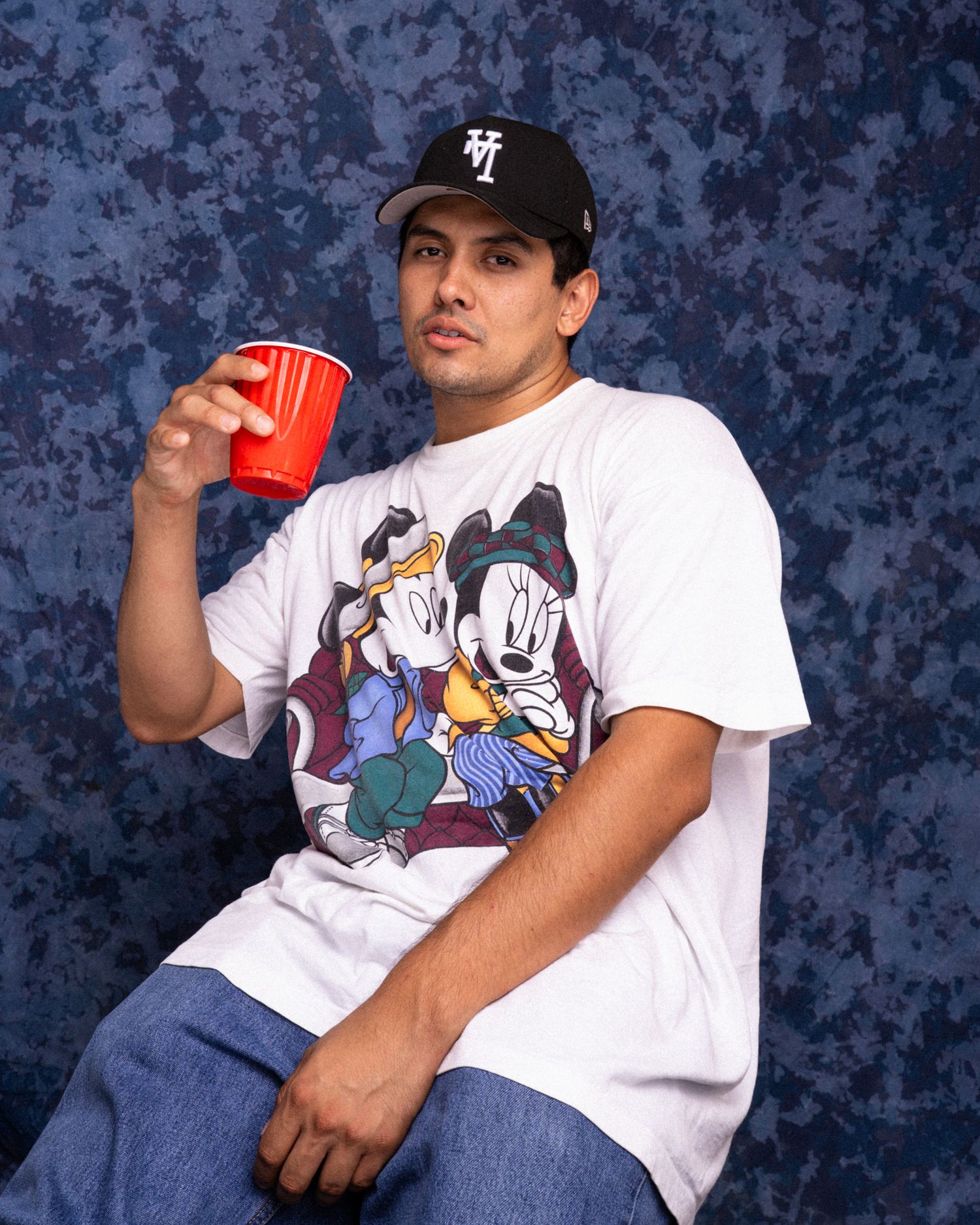 Man in black cap and Mickey Mouse tee holds red cup, posed against blue mottled backdrop.