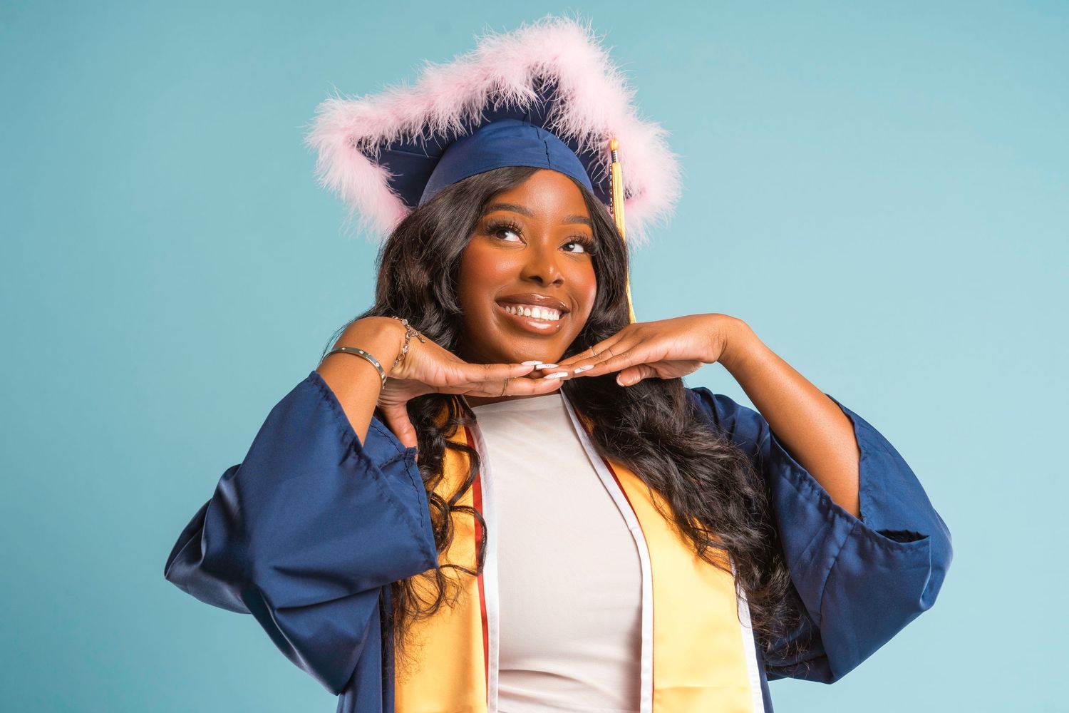 Smiling young woman in blue graduation gown and cap with pink fur trim posing against light blue background.