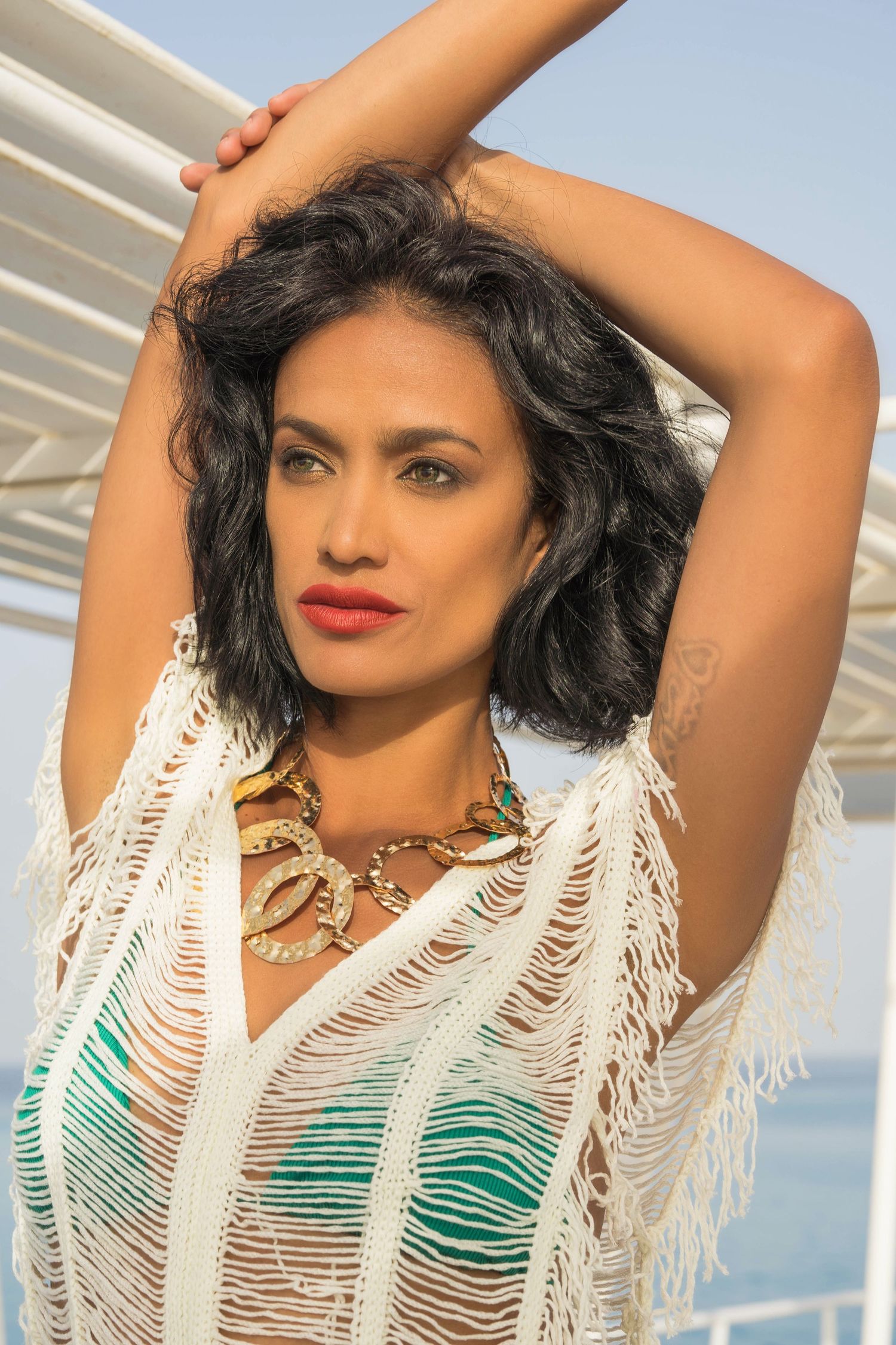 Fashion model wearing fringed beach cover-up and statement necklace poses against ocean view.
