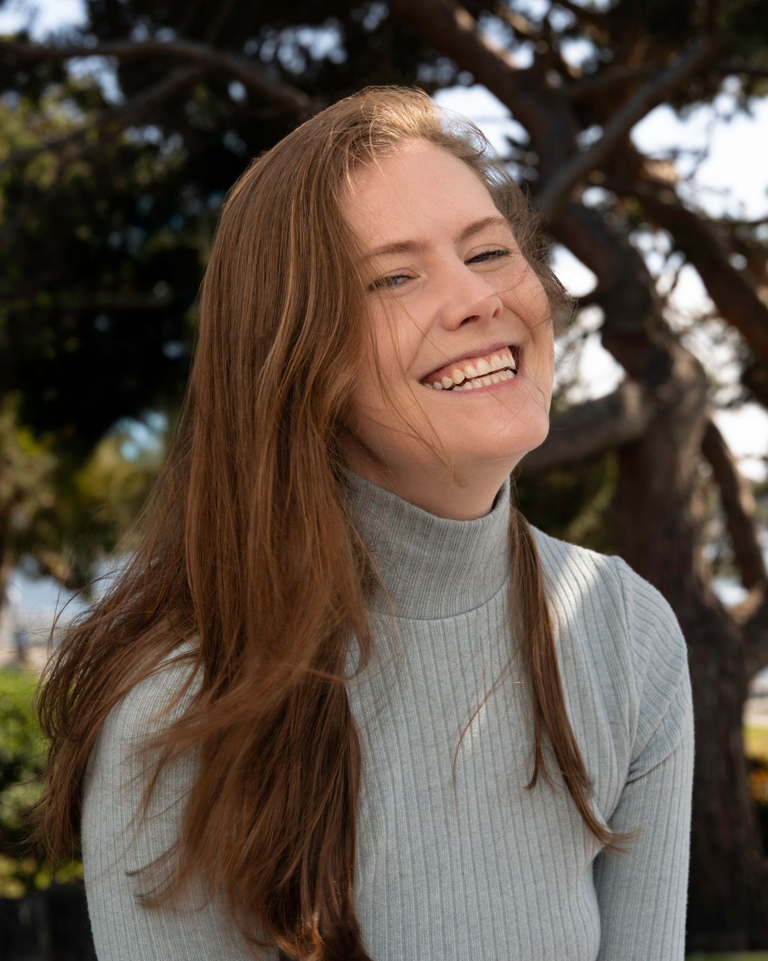 A person in a gray turtleneck sweater laughs joyfully while their hair flows in the wind.