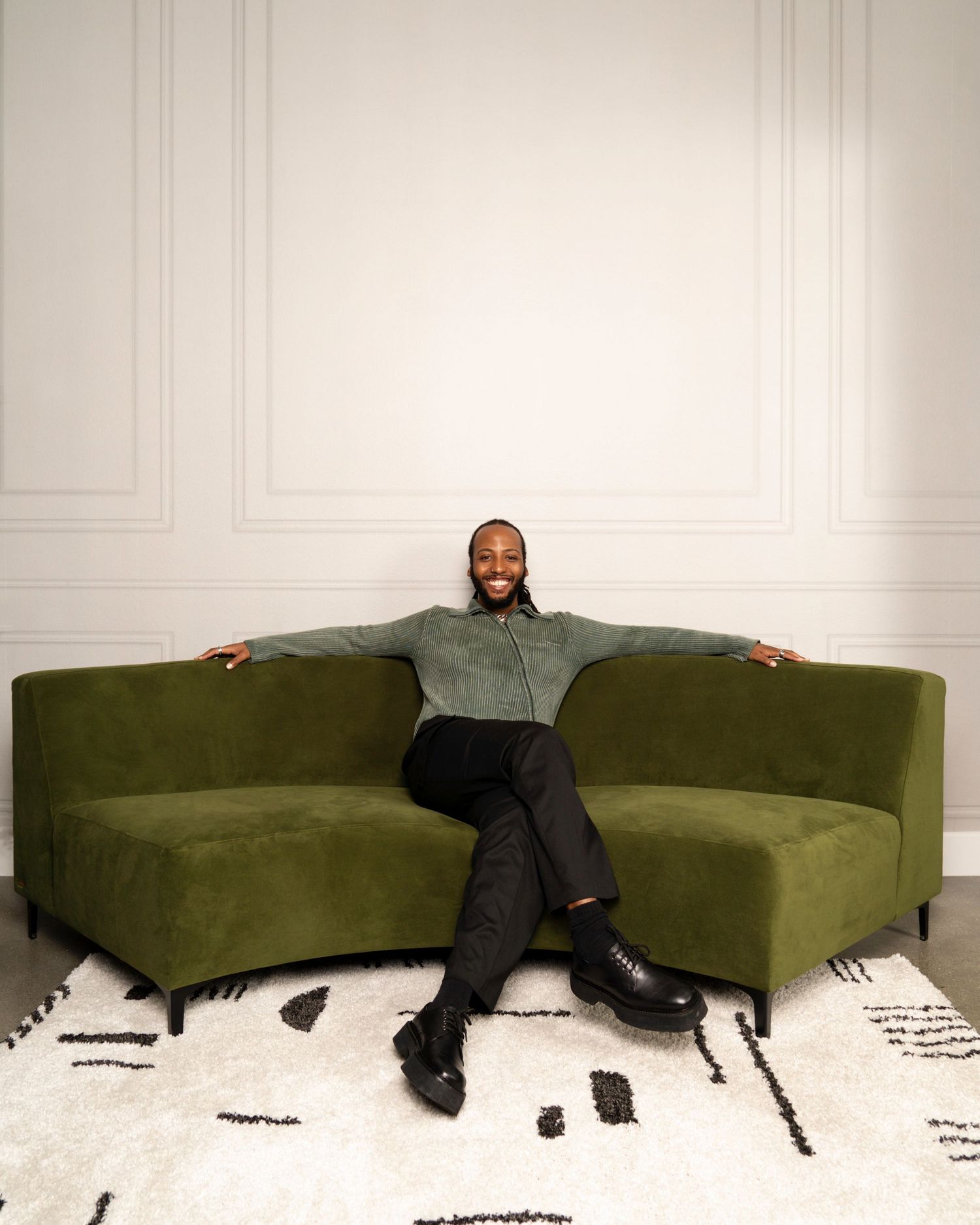 A person lounges on an olive green couch against a white wall with a black and white patterned rug below.