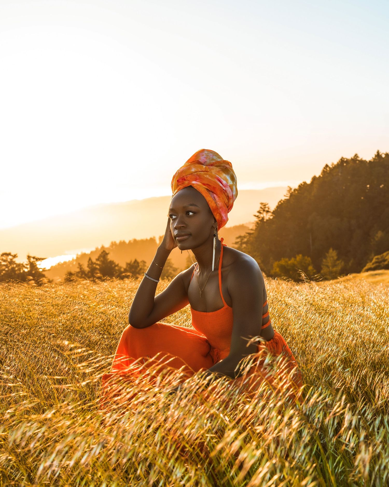 Person in orange headwrap and tank top poses thoughtfully in golden mountain field.