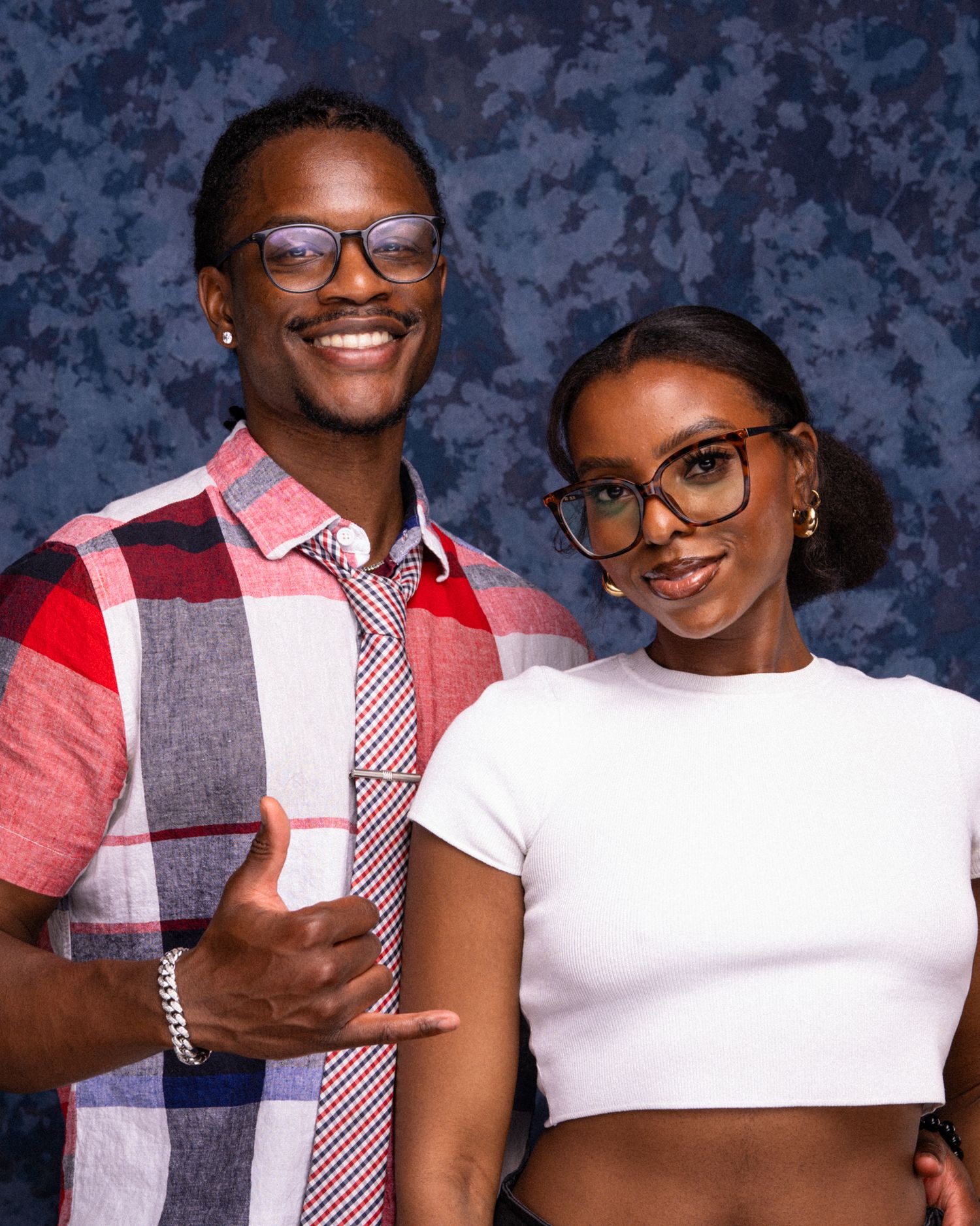 Two stylish young adults wearing glasses pose together, man in plaid shirt giving thumbs up, woman in white crop top.