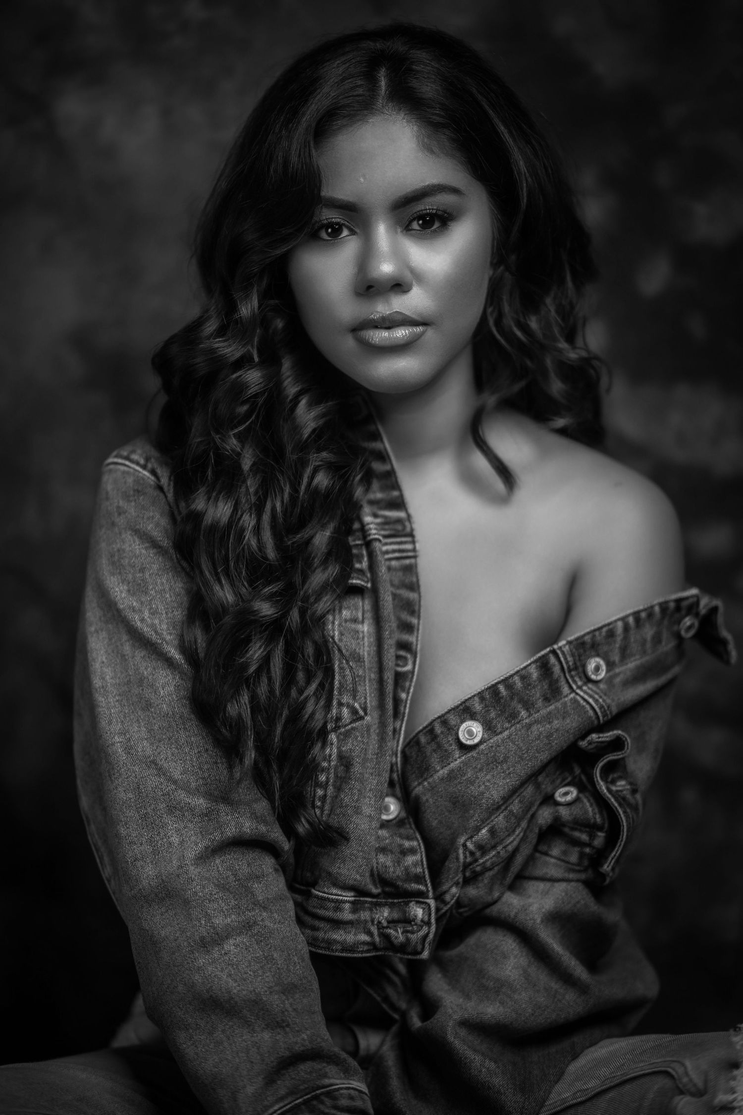 Black and white portrait of a young woman with long curly hair wearing an off-shoulder denim jacket.
