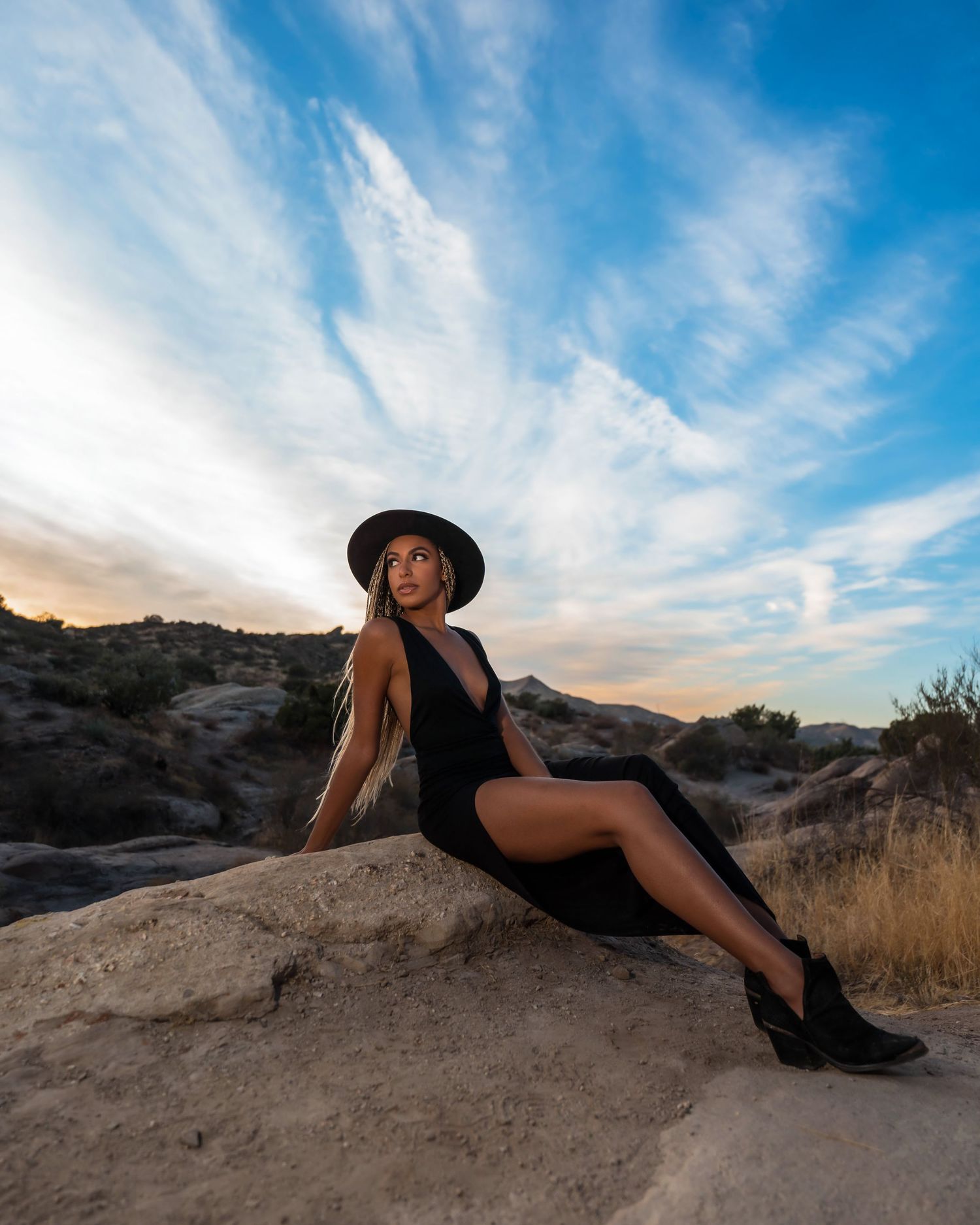 Woman in black dress and hat poses on desert rocks under a dramatic blue sky at sunset.