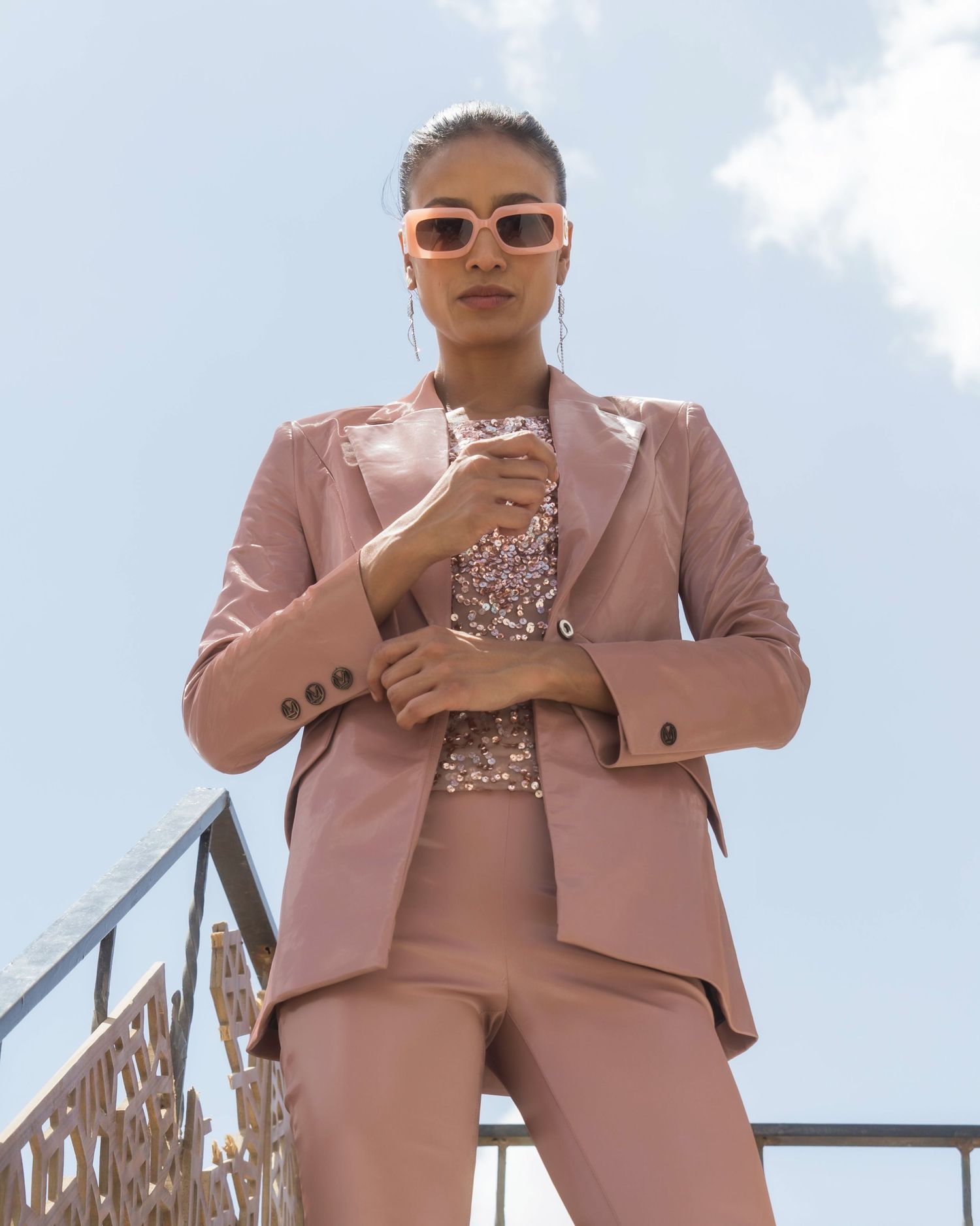 Model in pink pantsuit and sunglasses poses confidently against blue sky.