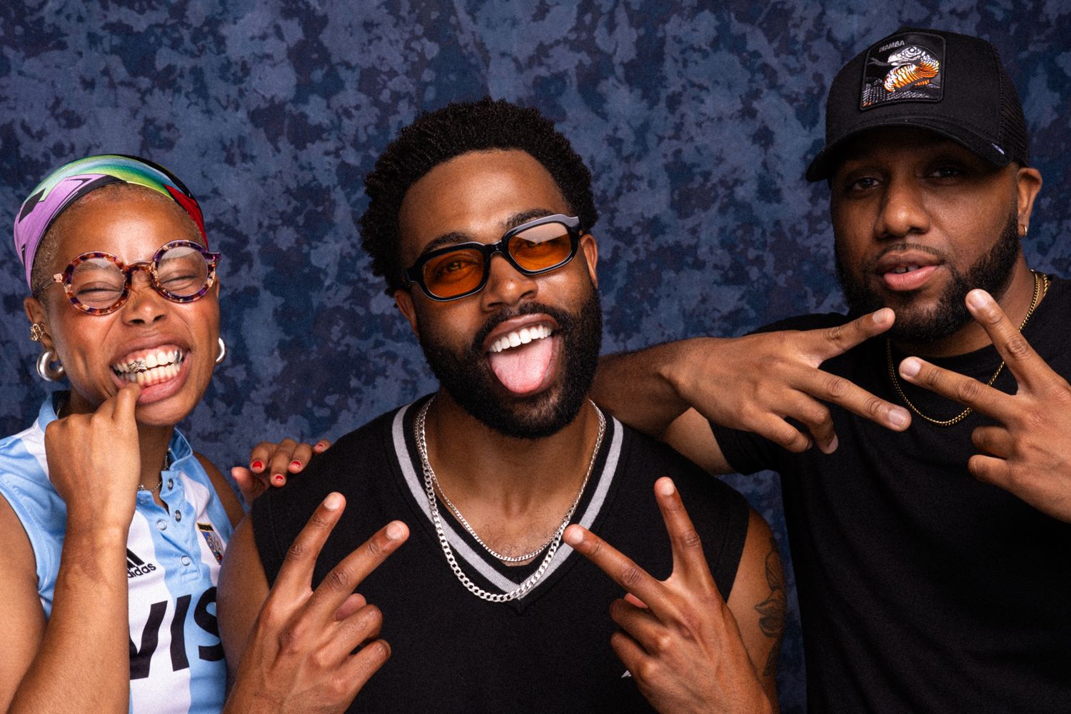 Three friends strike fun poses against a blue backdrop, with the center person sticking out their tongue.