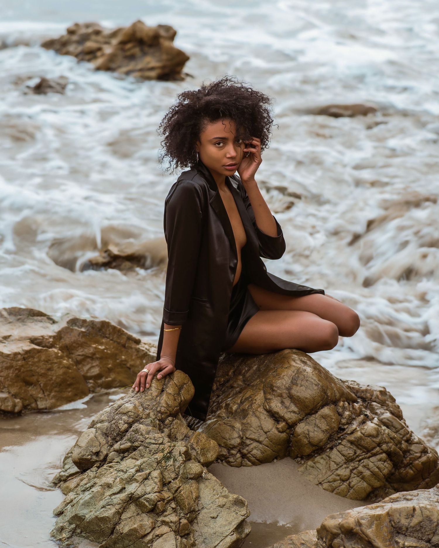 Young woman with curly hair in black outfit sitting on rocky beach with ocean waves in background.