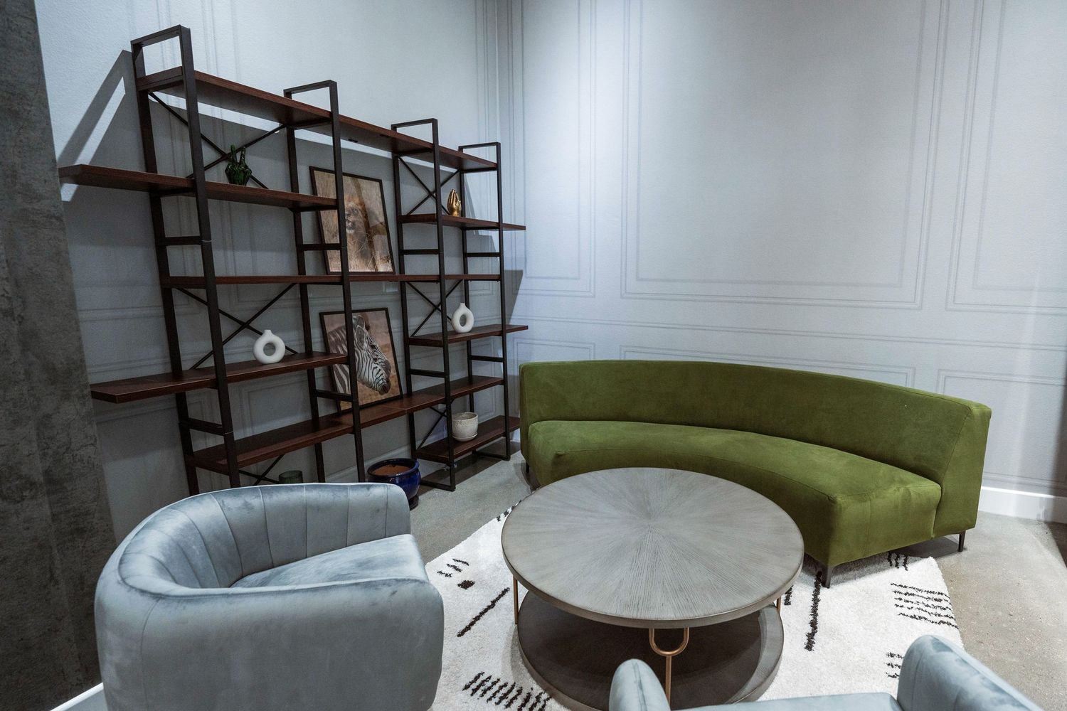 Modern lounge with curved green sofa, gray armchairs, round coffee table, and industrial wood bookshelf in elegant white room.