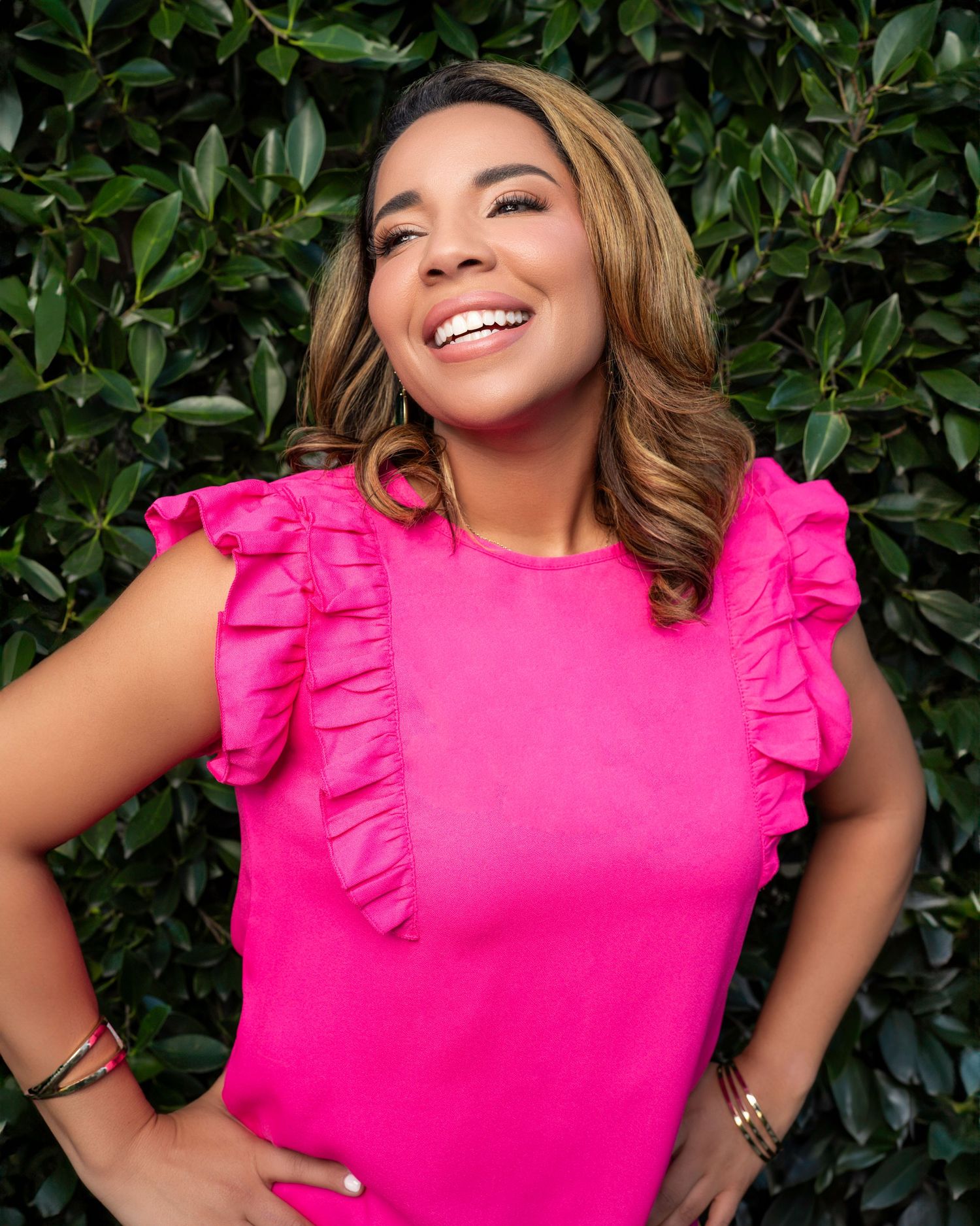 Woman in bright pink ruffled top smiles brightly against green leaf background.